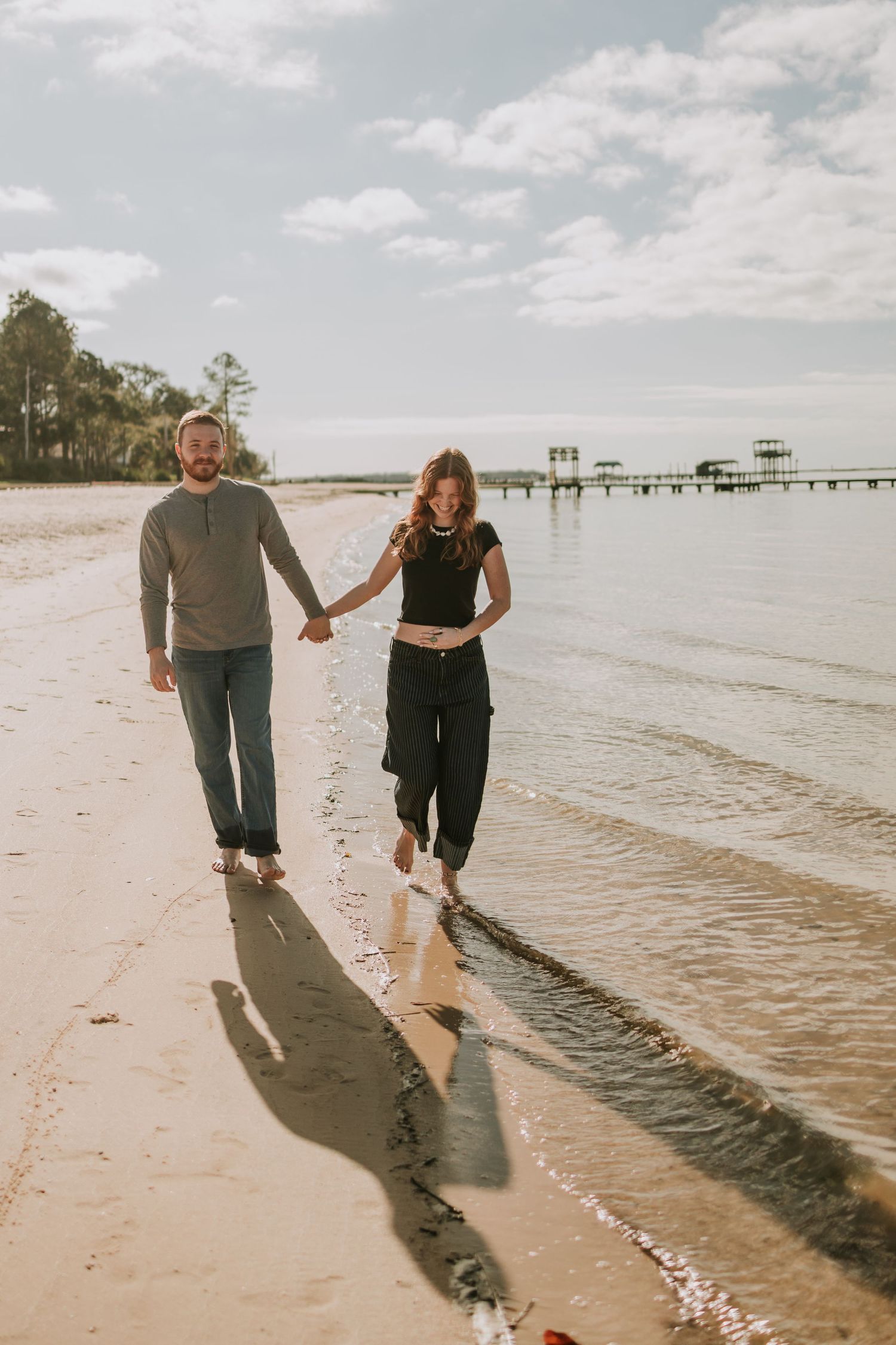 A couple walks hand-in-hand along a sandy beach shoreline, barefoot in shallow water on a sunny day.