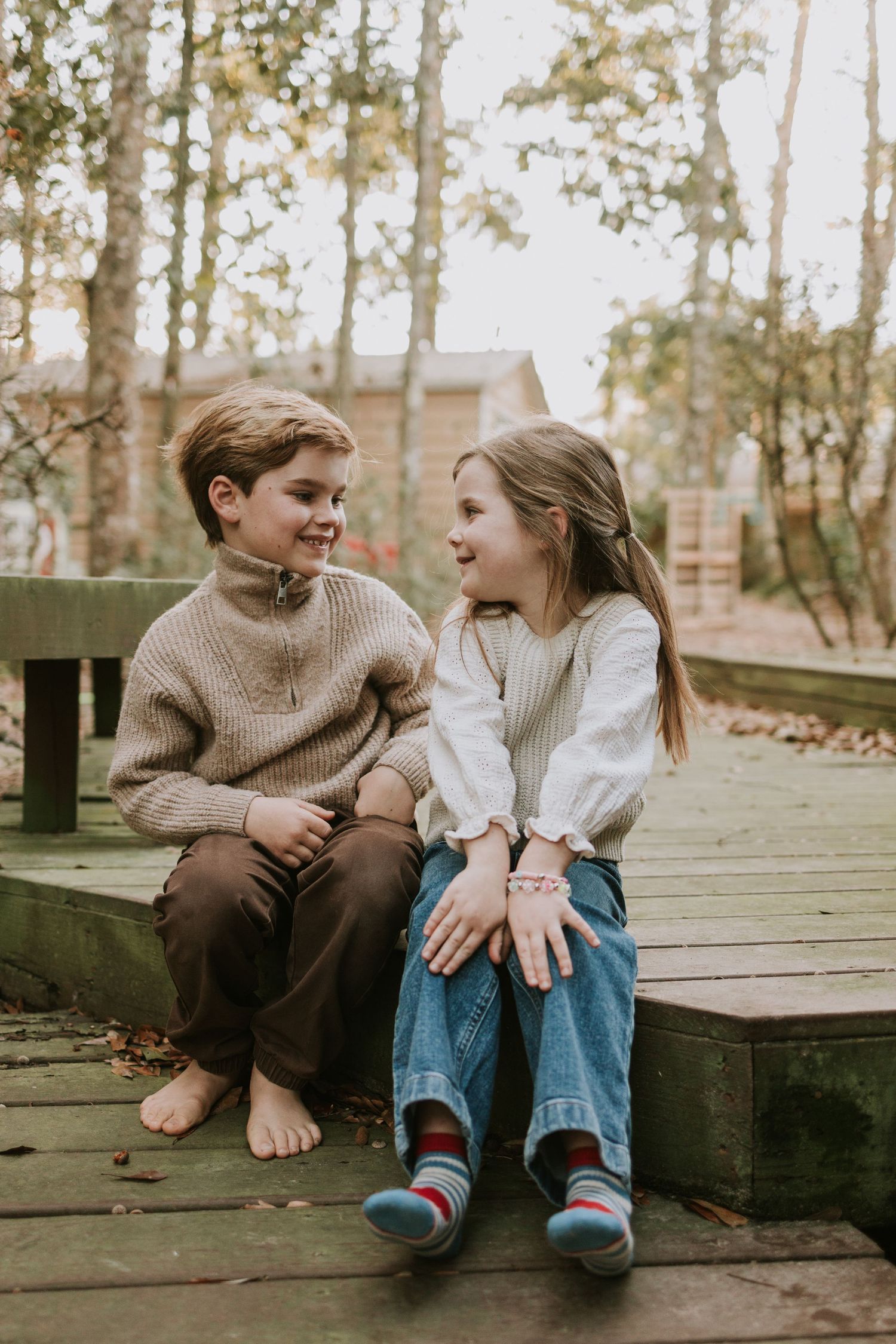 Two children sitting on a wooden deck in autumn, smiling at each other in cozy sweaters and colorful socks.