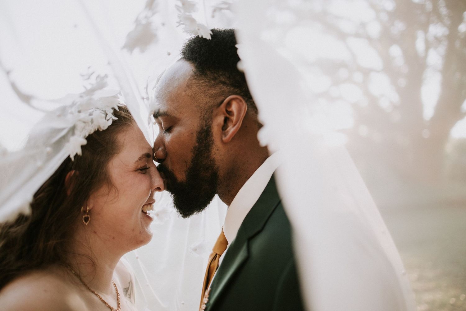 A bride and groom share a romantic moment under a veil, smiling and touching foreheads on their wedding day.