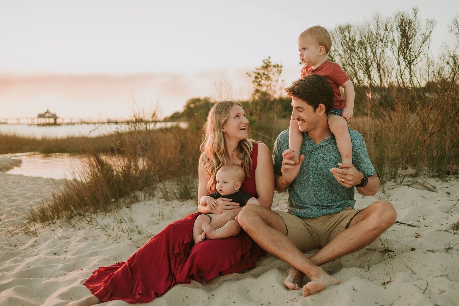 Happy family sitting on sandy beach at sunset, parents with two young children enjoying a warm golden hour moment.