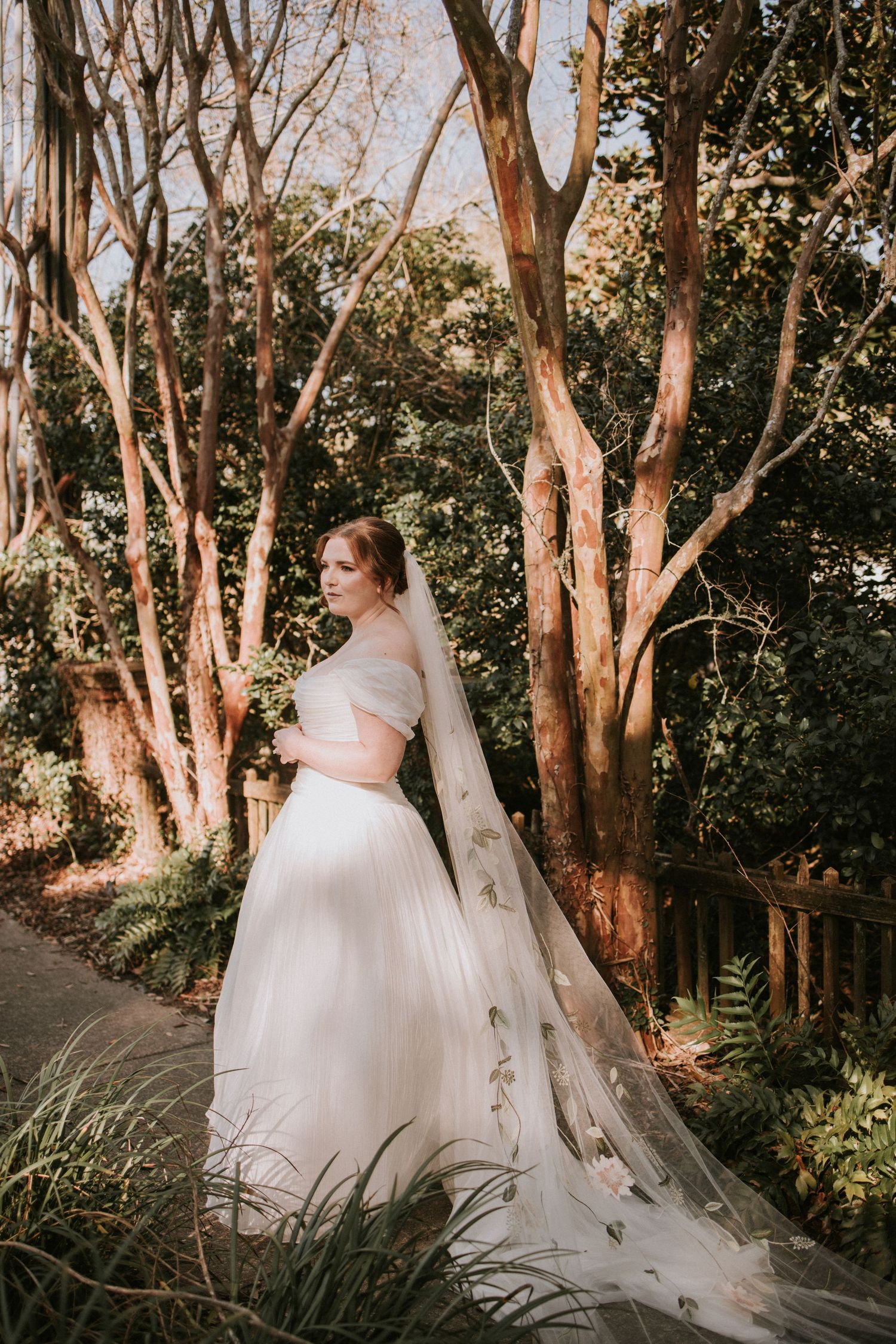 Bride in white off-shoulder gown with floral veil stands among sunlit trees in a lush garden setting.
