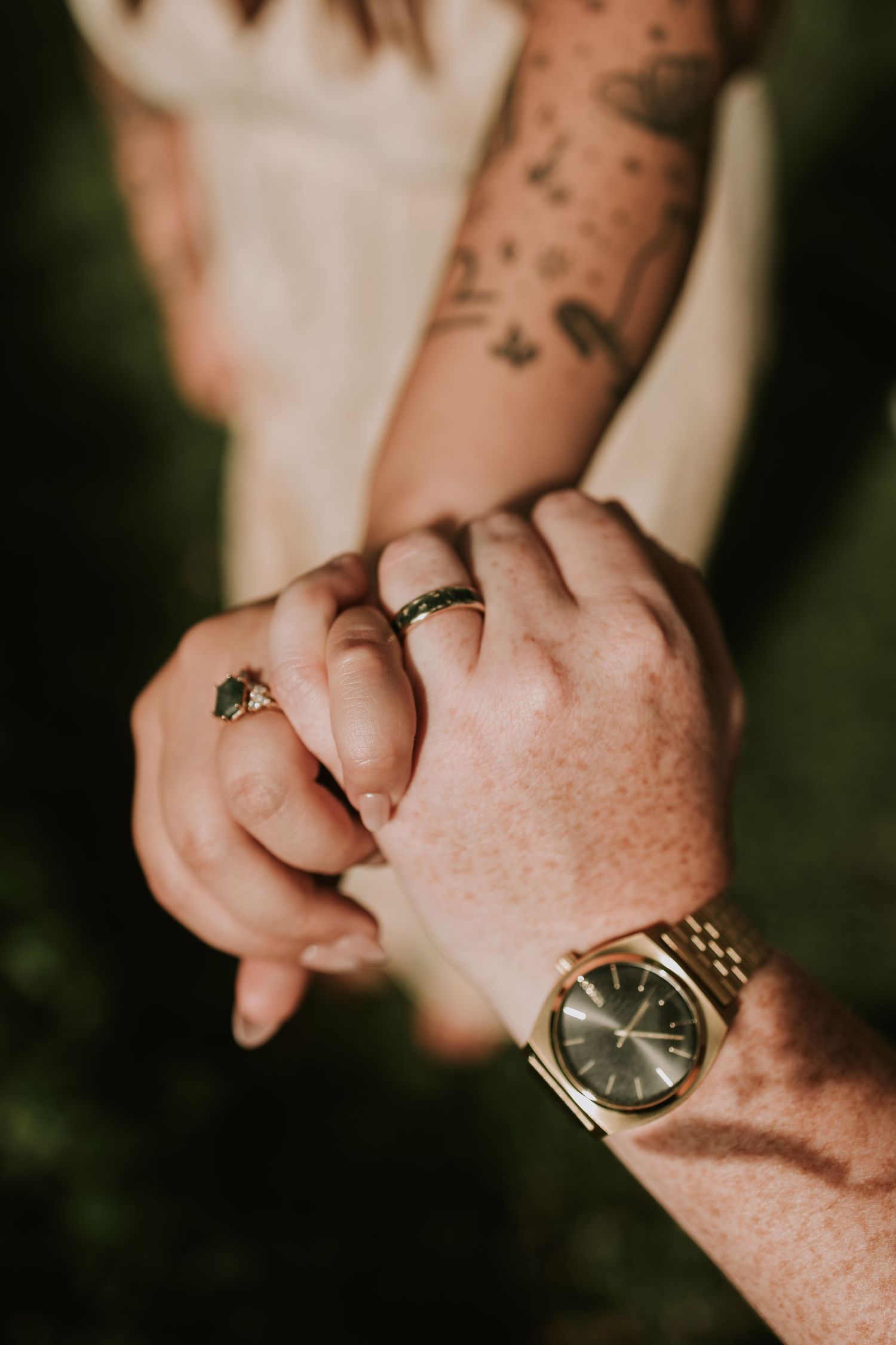 Two hands holding together, one with a gold watch and freckles, the other with tattoos and rings, symbolizing love.