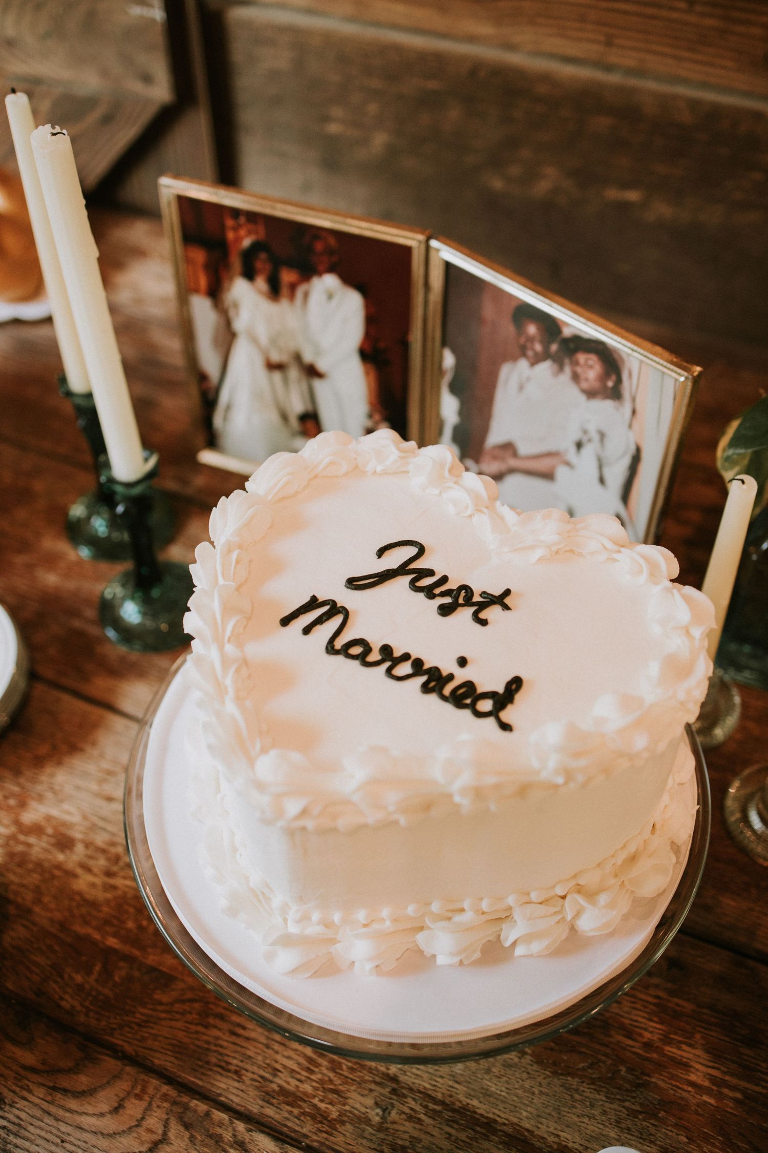 Heart-shaped white wedding cake with Just Married script, flanked by candles and vintage couple photos on rustic wood table.