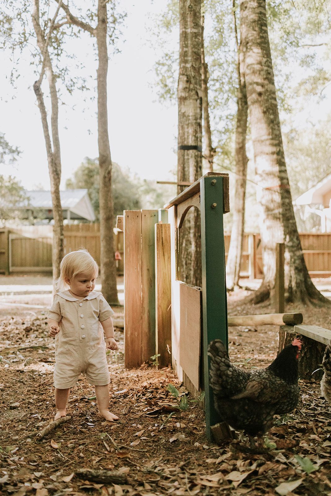 Small child in light clothing exploring outdoors near wooden fencing and trees.