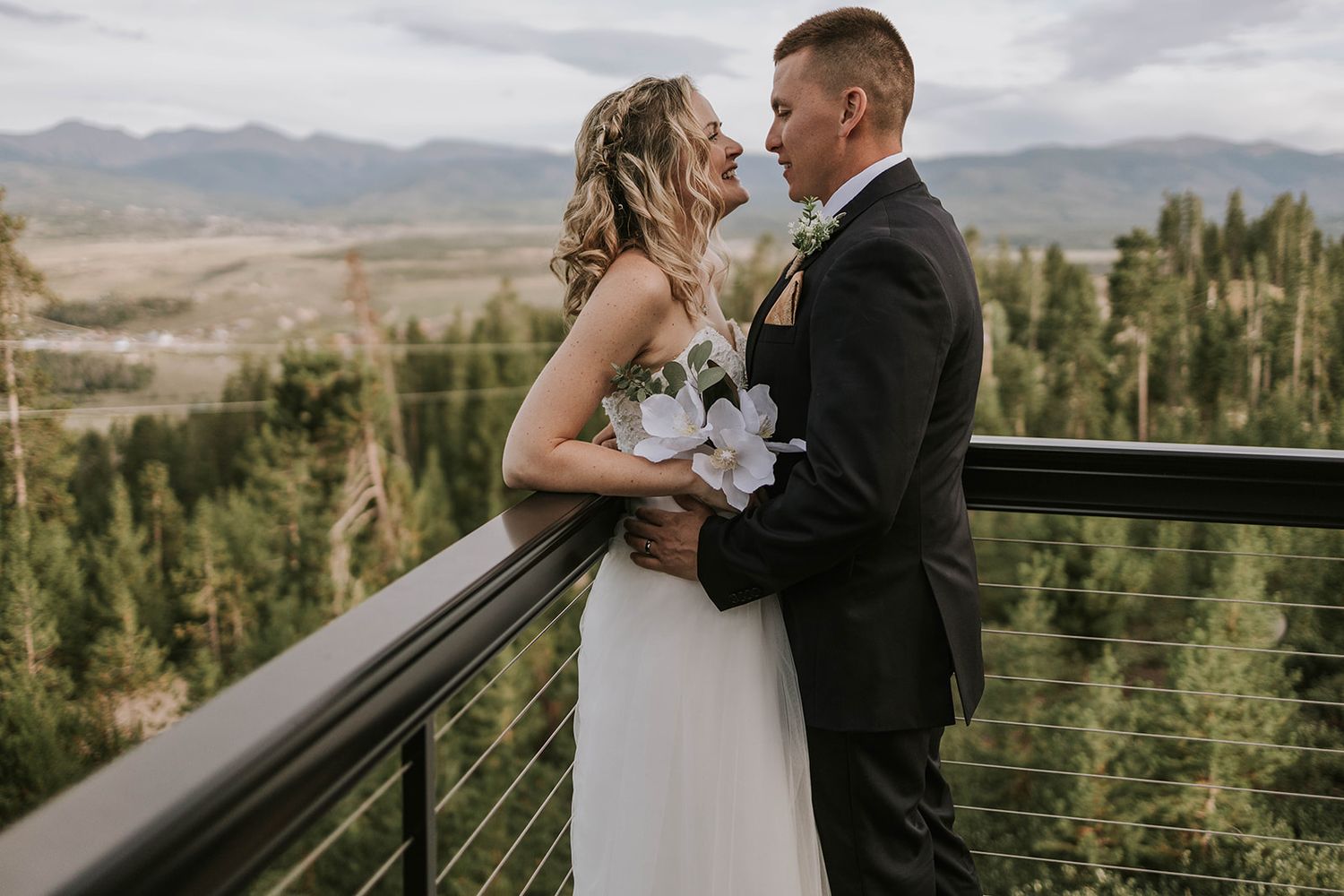 A couple shares an intimate moment on a mountain deck overlooking scenic forest views while dressed in wedding attire.