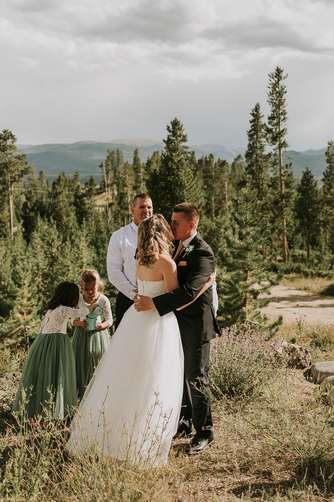 A small wedding ceremony takes place in a scenic mountain forest setting with pine trees and natural landscape.