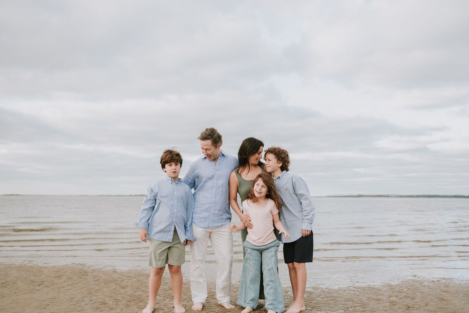 Family of five posing together on a sandy beach with calm ocean waters and overcast sky in the background.