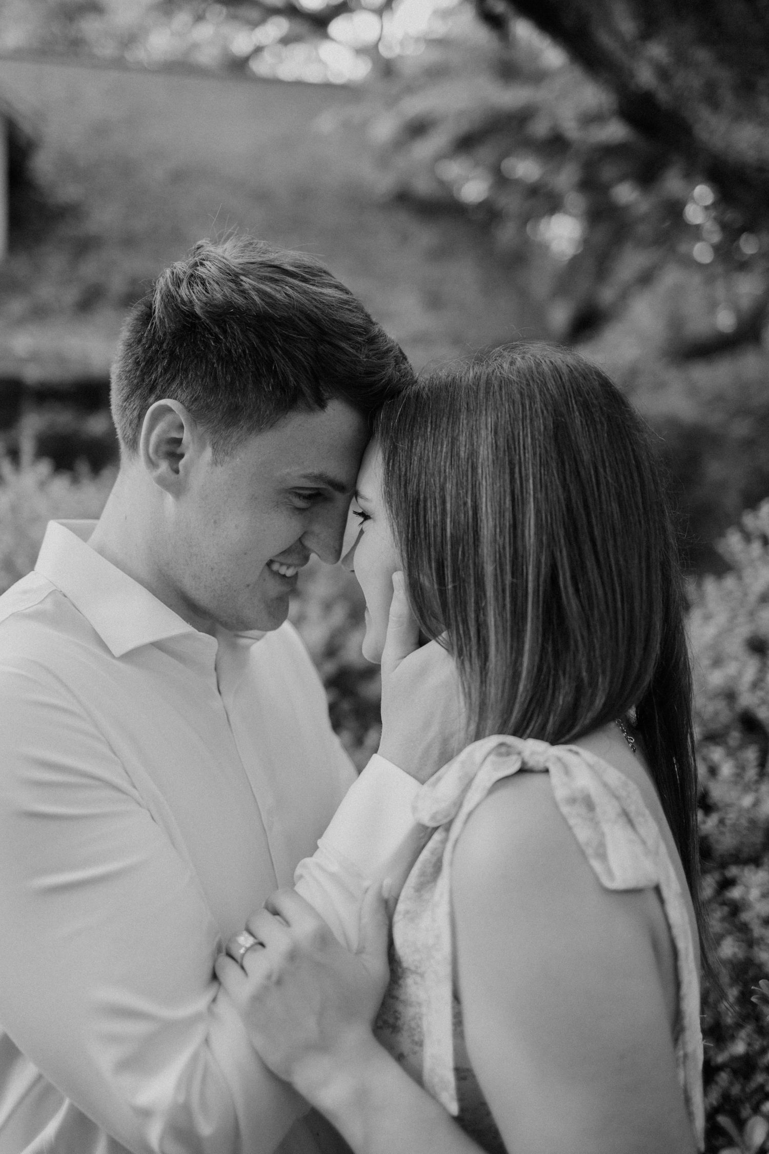 Black and white photo of a smiling couple touching foreheads in a romantic outdoor garden setting.