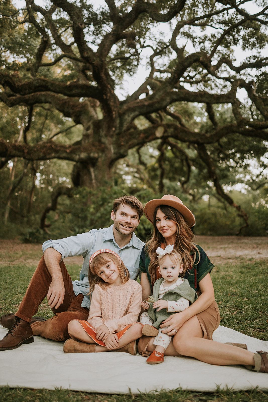 Family sits together on white blanket under sprawling oak tree branches in outdoor portrait setting.