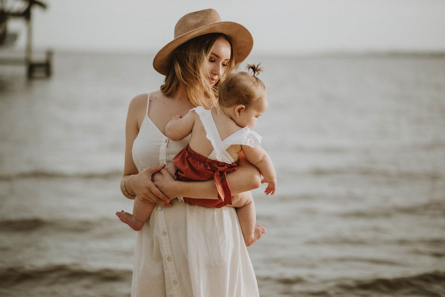 Figure in a white dress and brown hat holding small child by the water at sunset.