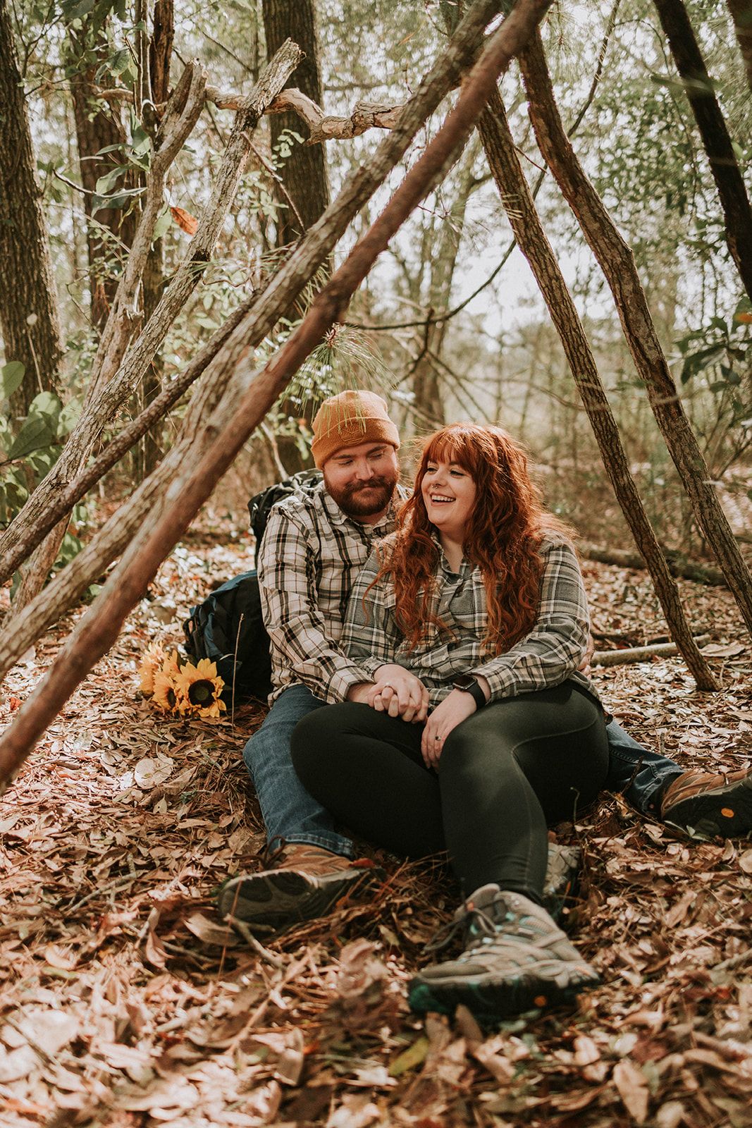A couple in matching plaid shirts sits together on fallen leaves in a wooded forest setting during autumn.
