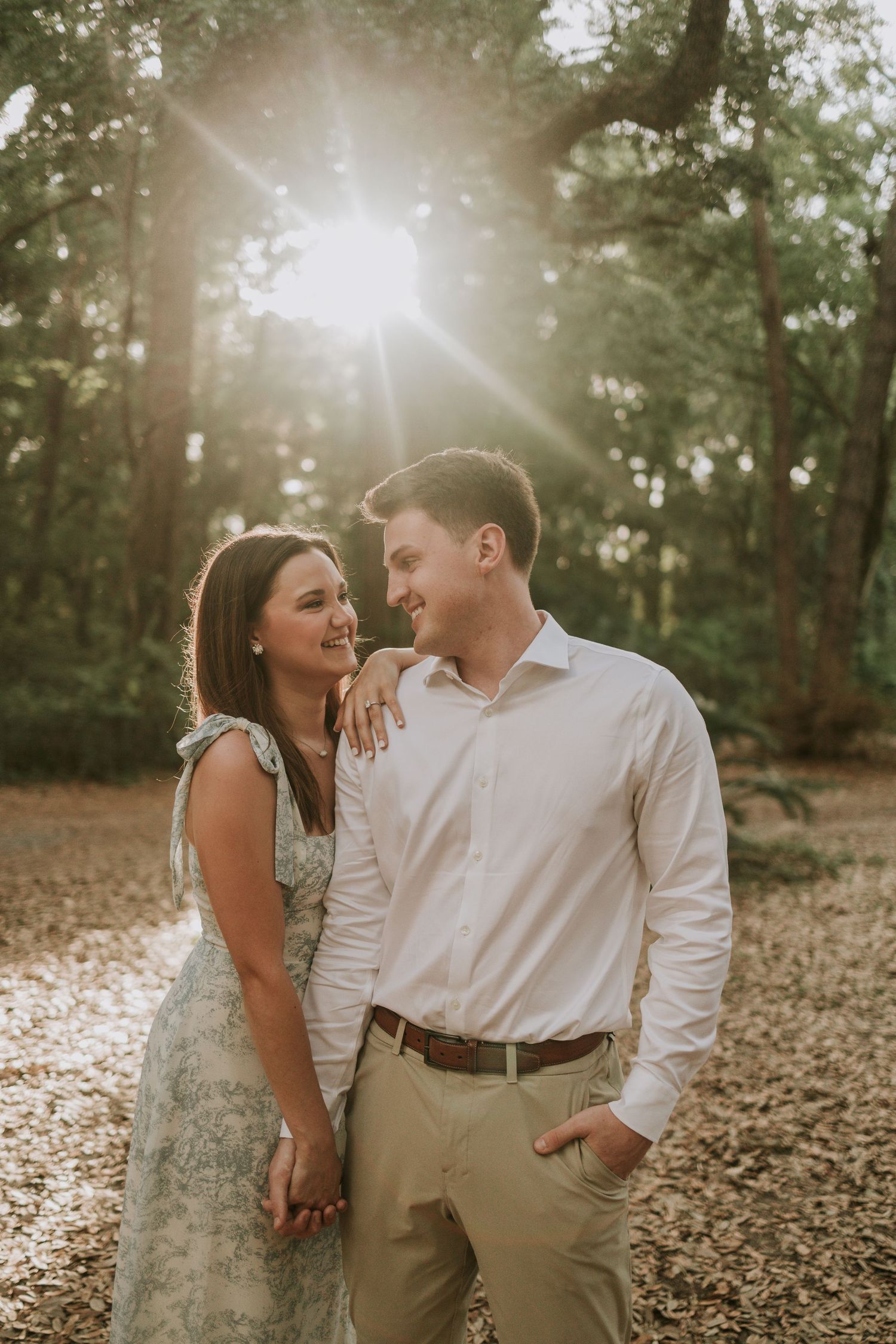 Couple holding hands and smiling at each other in a sunlit forest during golden hour engagement photo.