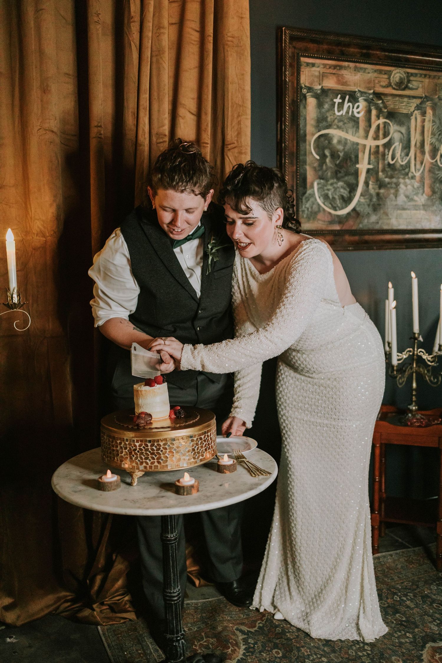 Two brides cutting their wedding cake together at an elegant venue with candlelight and vintage decor.