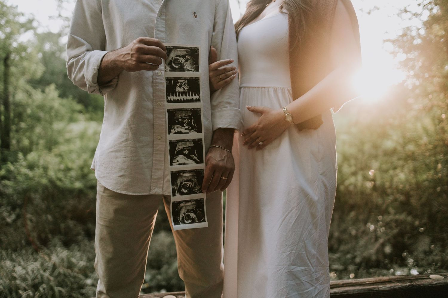 Couple holding ultrasound photos outdoors at sunset, woman in white dress with baby bump, maternity announcement photo.
