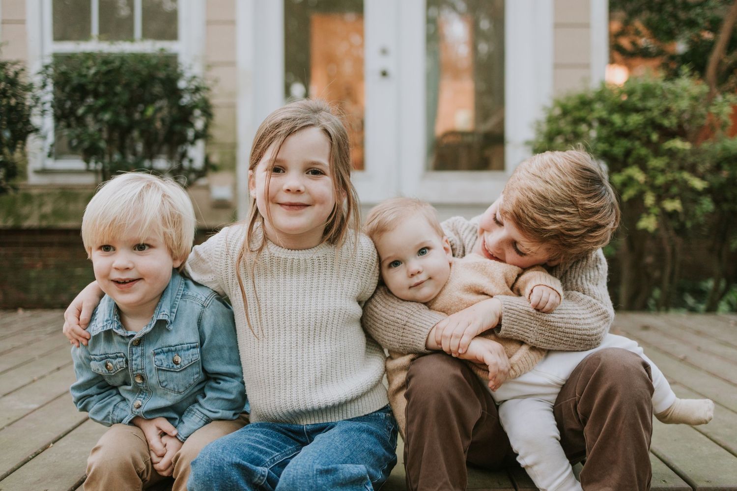 Four young siblings smiling and hugging together outdoors on a wooden deck in front of a house.