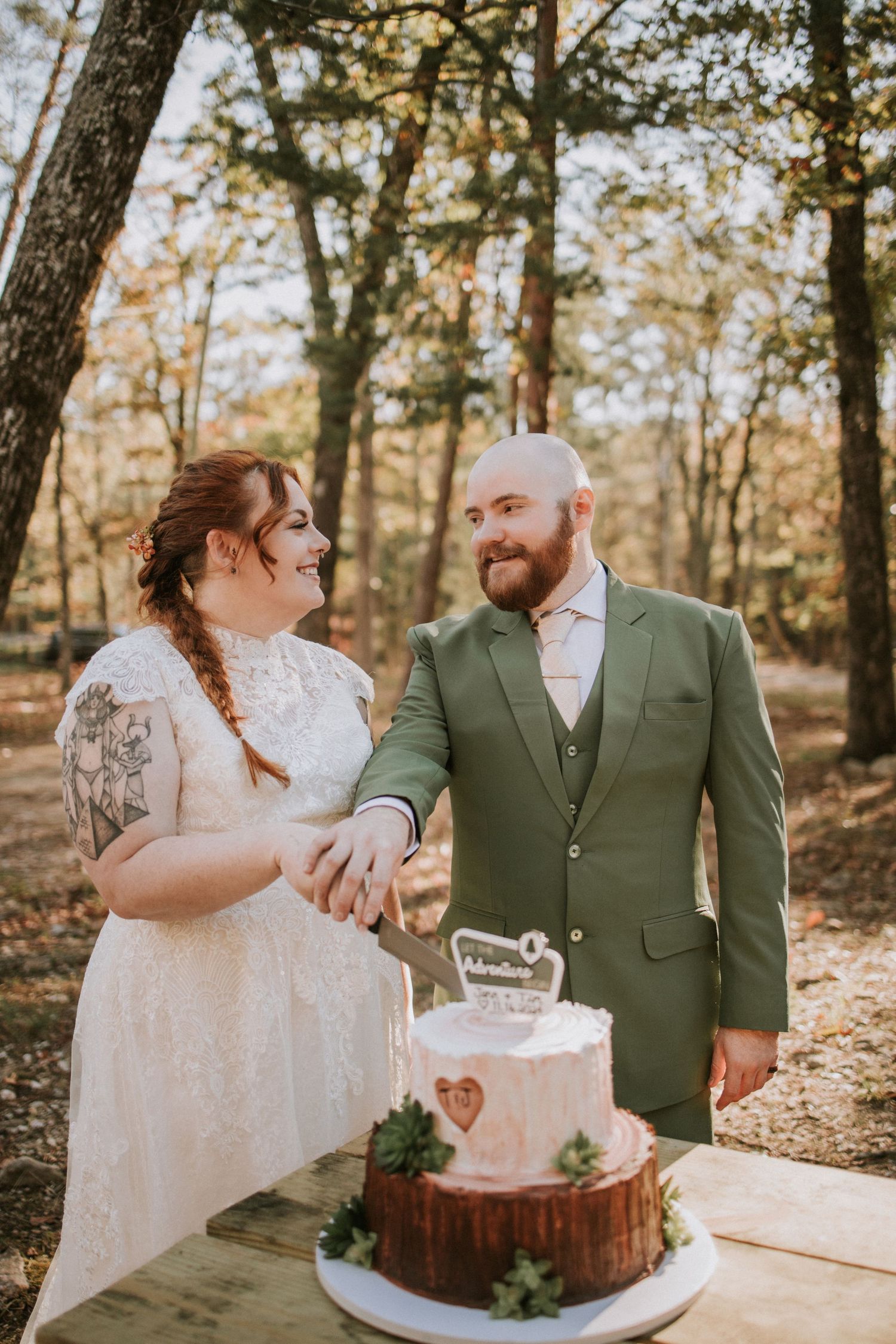 Newlyweds in forest setting prepare to cut their elegant pink wedding cake displayed on rustic wood slice.