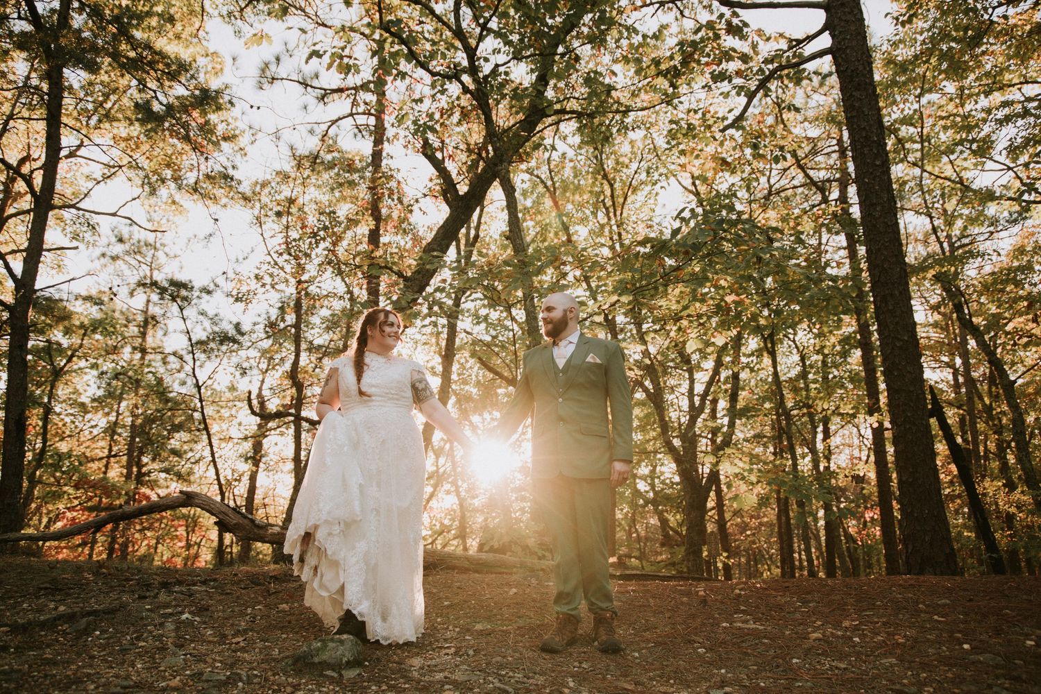A couple holds hands walking through a sunlit autumn forest during their outdoor wedding ceremony.