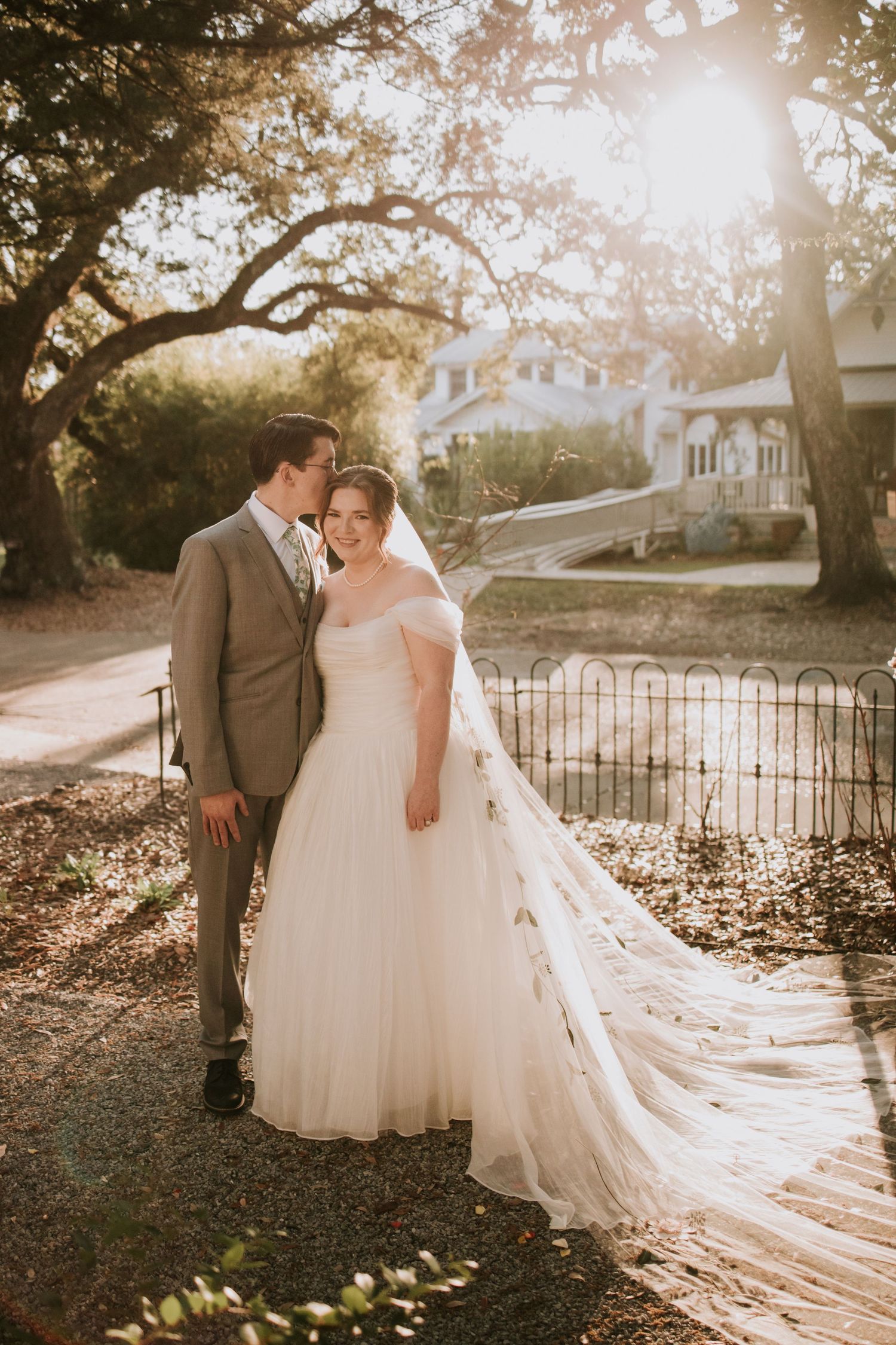 Bride and groom pose in golden sunlight, her flowing white gown and veil trailing elegantly on a tree-lined path.