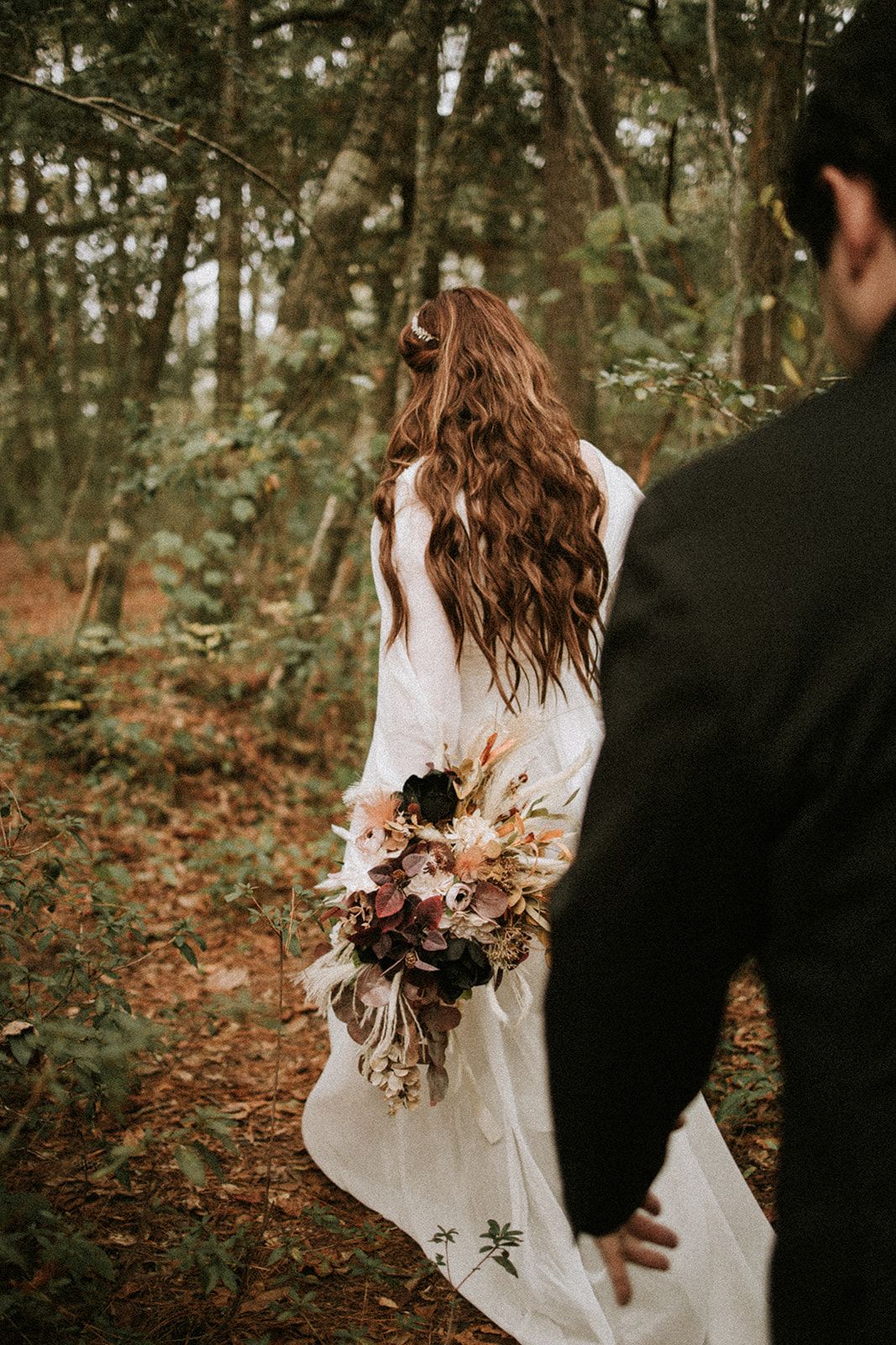 Bride in white dress holds dried flower bouquet while walking through autumn forest with partner.