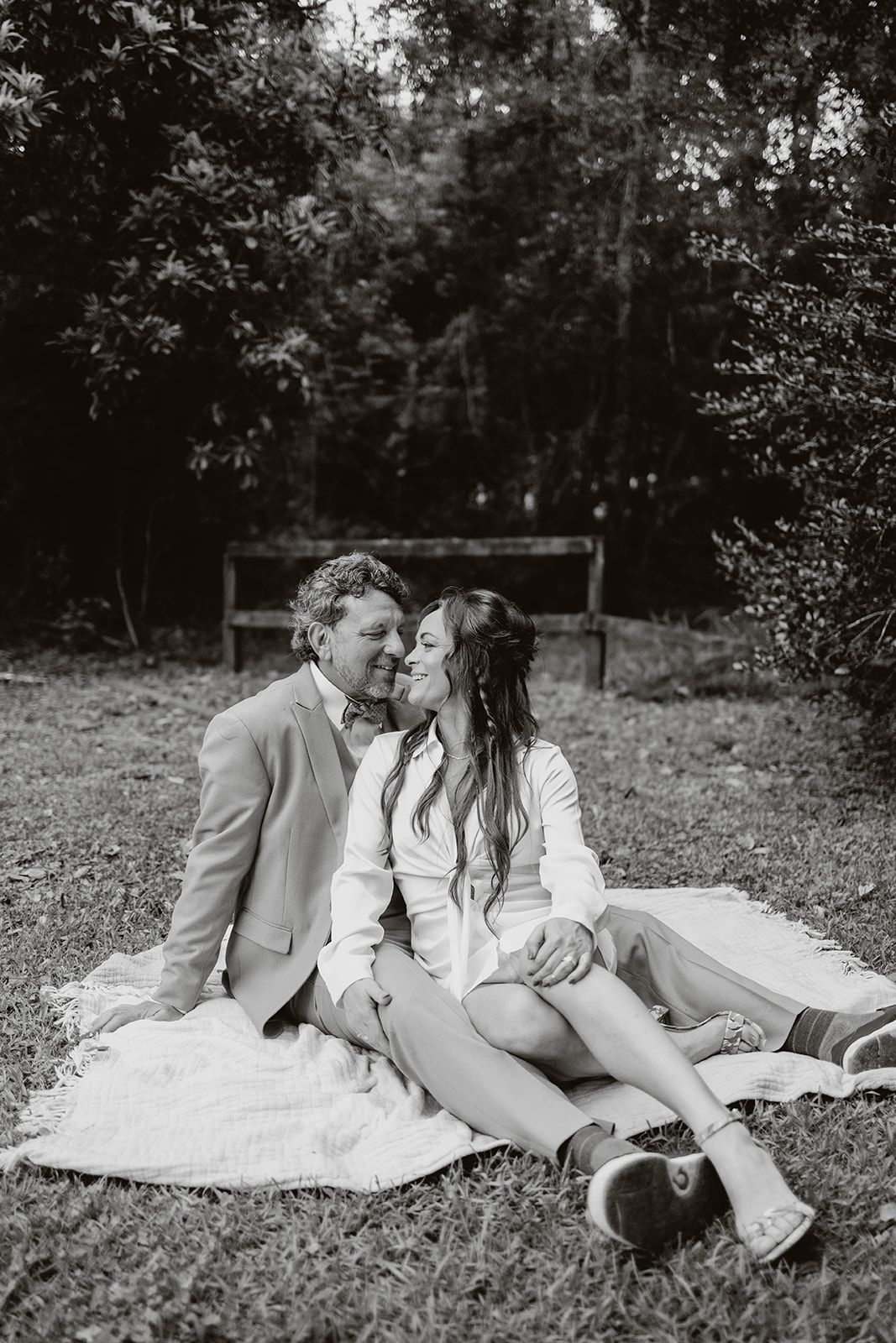 Romantic couple shares tender moment on picnic blanket in wooded setting, captured in black and white photography.