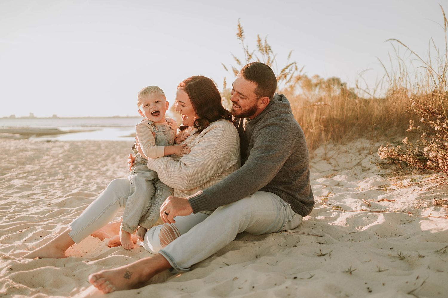 A couple and young child sit together during sunset at the beach with warm golden lighting in the background.