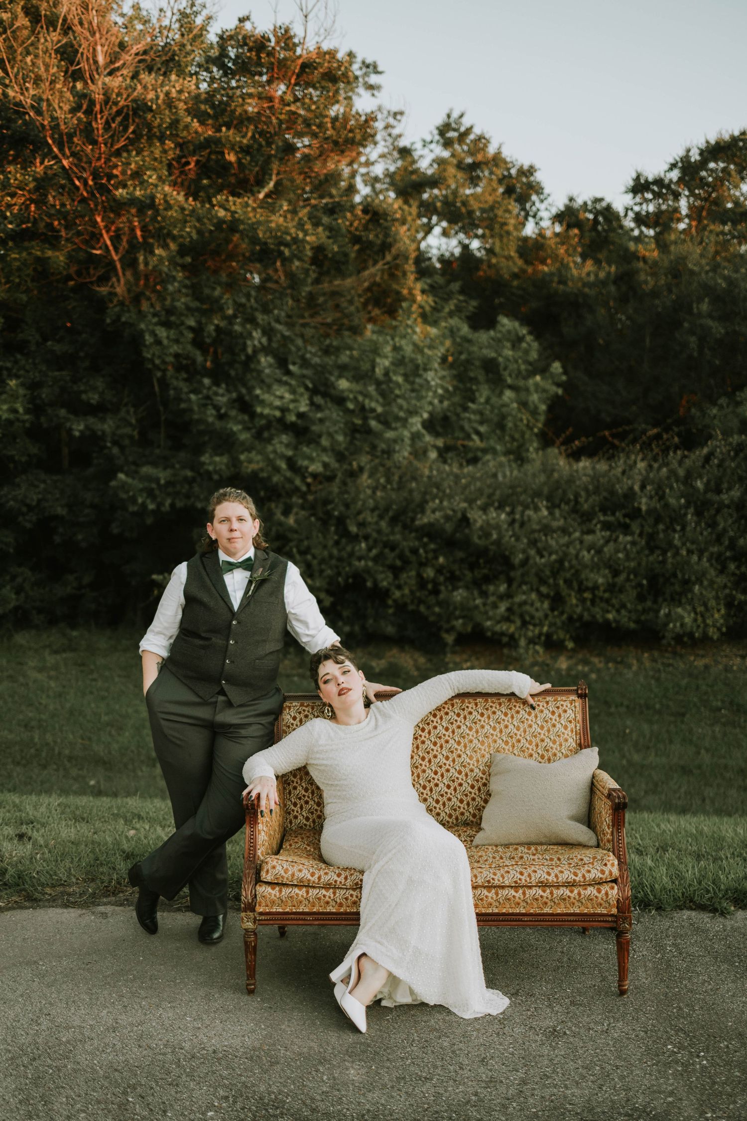 Couple poses outdoors on vintage gold settee in wedding attire, surrounded by lush green trees at golden hour.