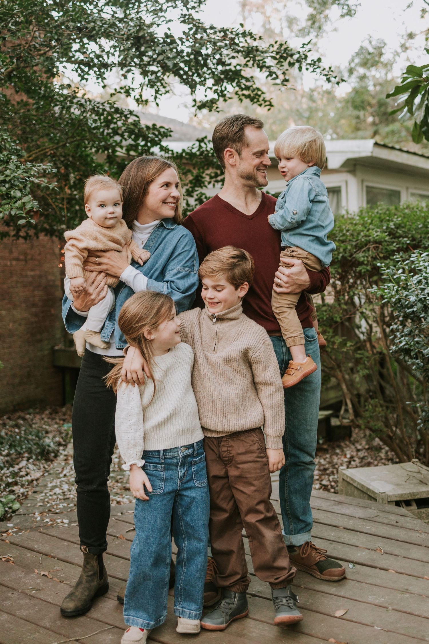 Happy family of six posing outdoors on a wooden deck surrounded by lush greenery, parents holding toddlers with children smiling.