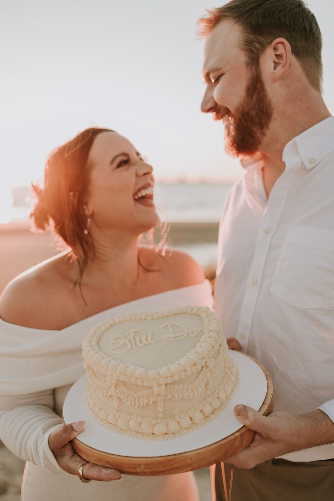 A couple laughs together while holding a decorative white cake at sunset during an outdoor celebration.