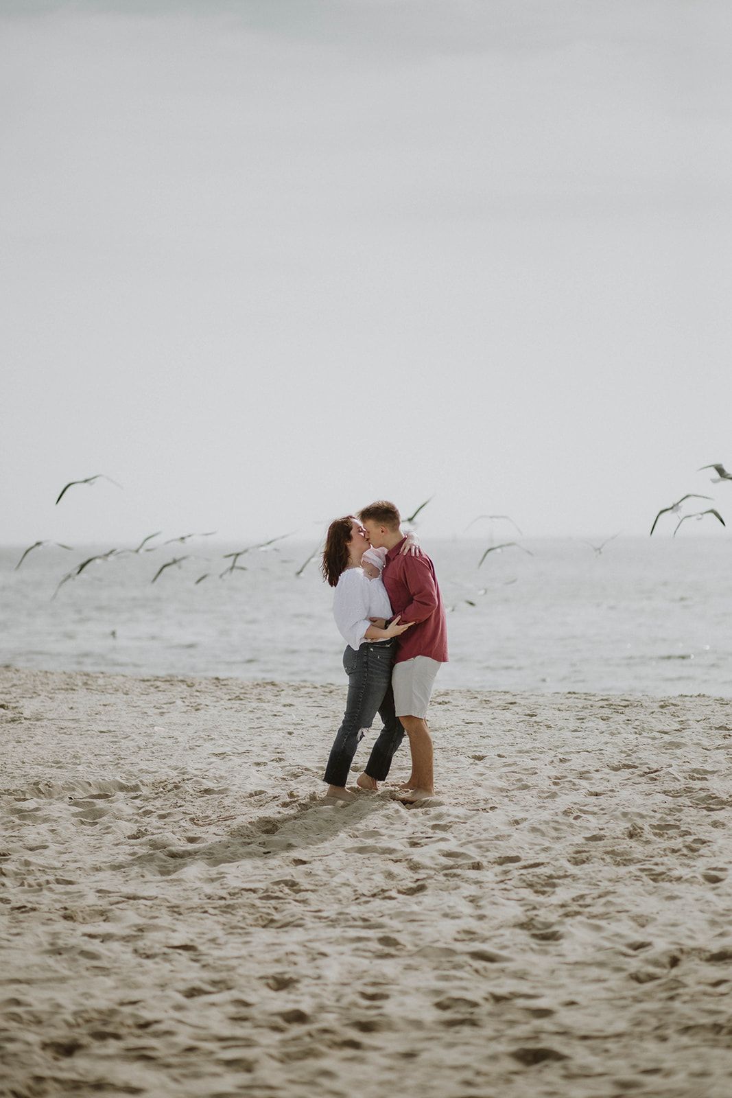 A couple shares a tender moment on a misty beach with seabirds flying overhead.