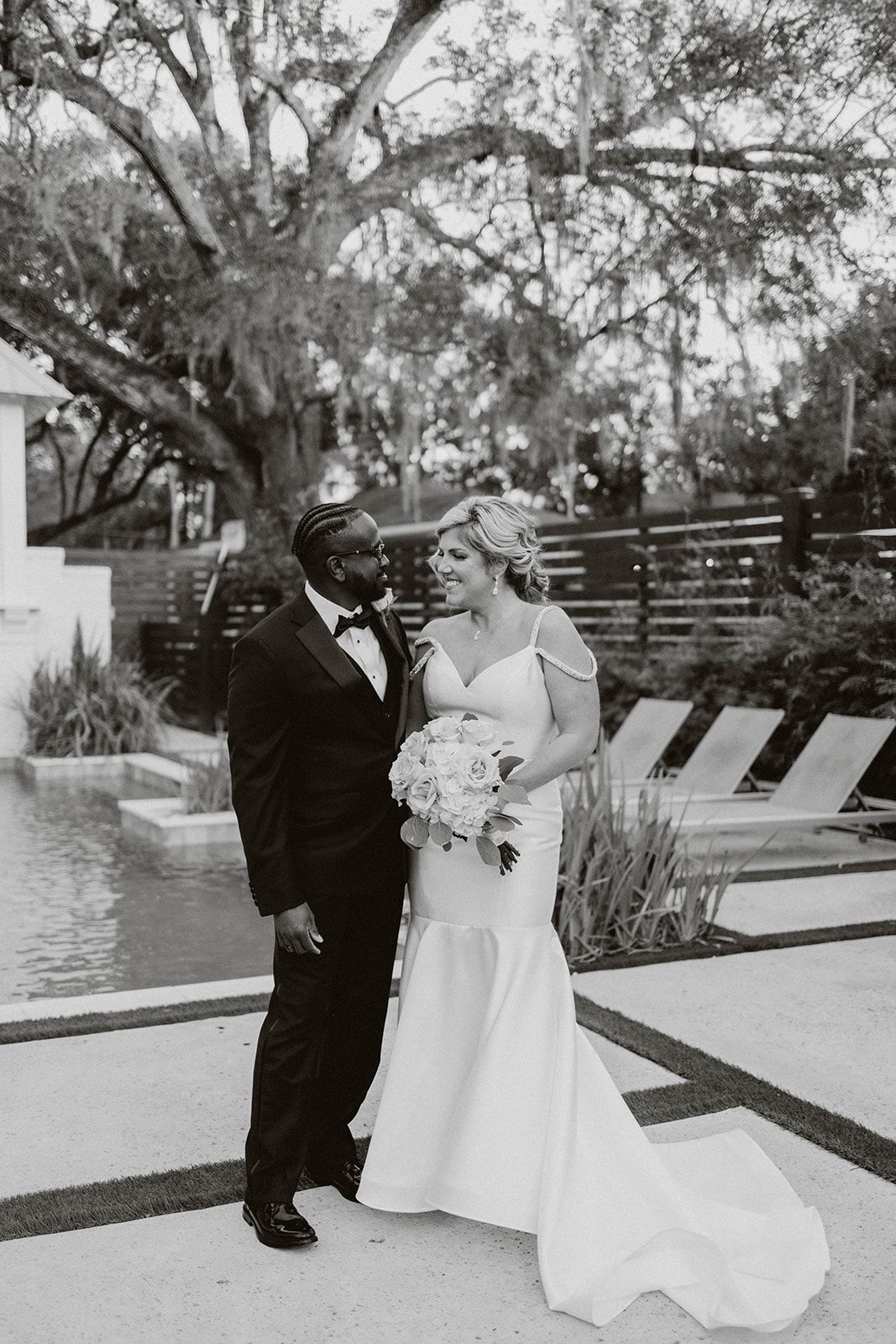 Wedding couple shares an intimate moment while standing by a pool surrounded by trees in black and white photography.