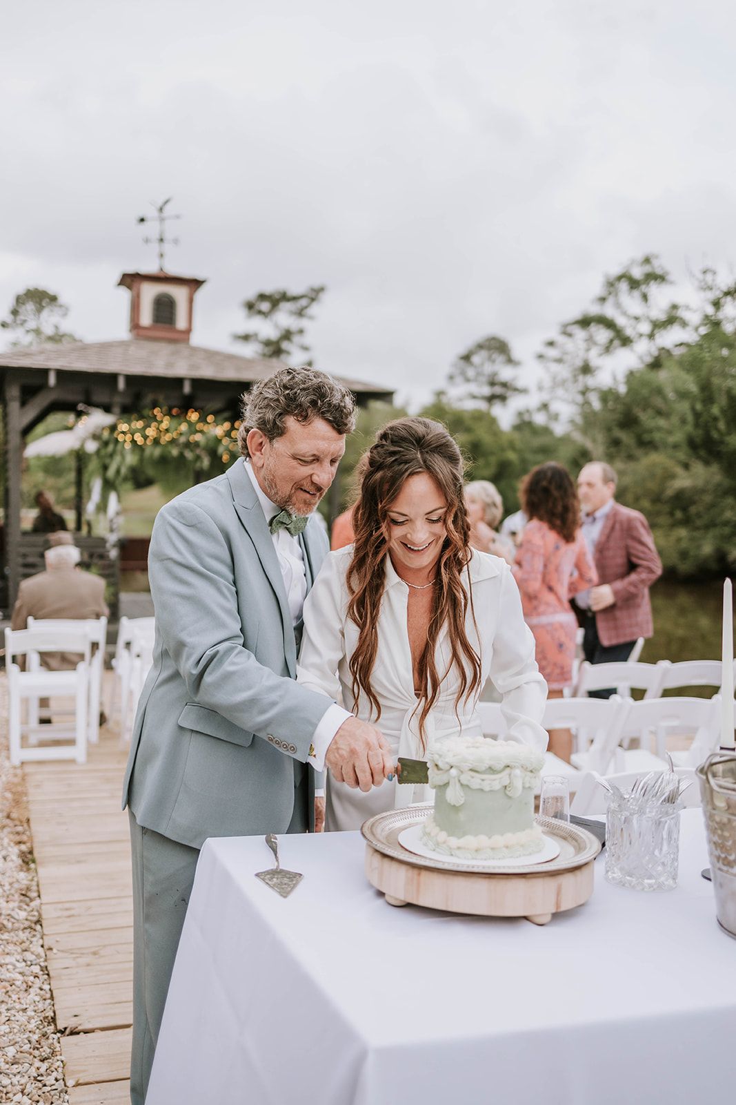 A couple cuts their elegant sage green wedding cake together at an outdoor venue with a rustic gazebo in the background.