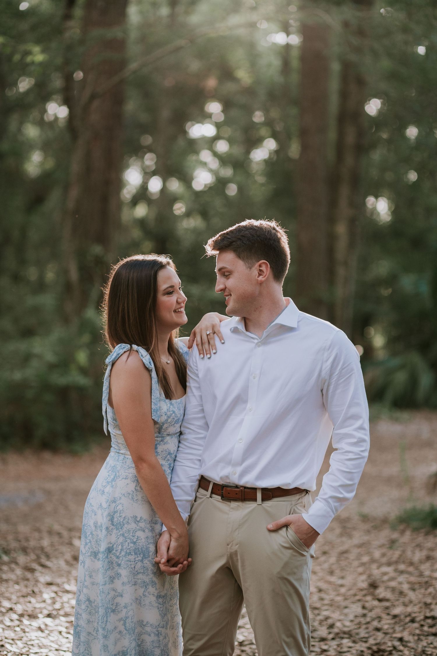 Couple holding hands and smiling at each other in a sunlit forest, woman in floral dress, man in white shirt.