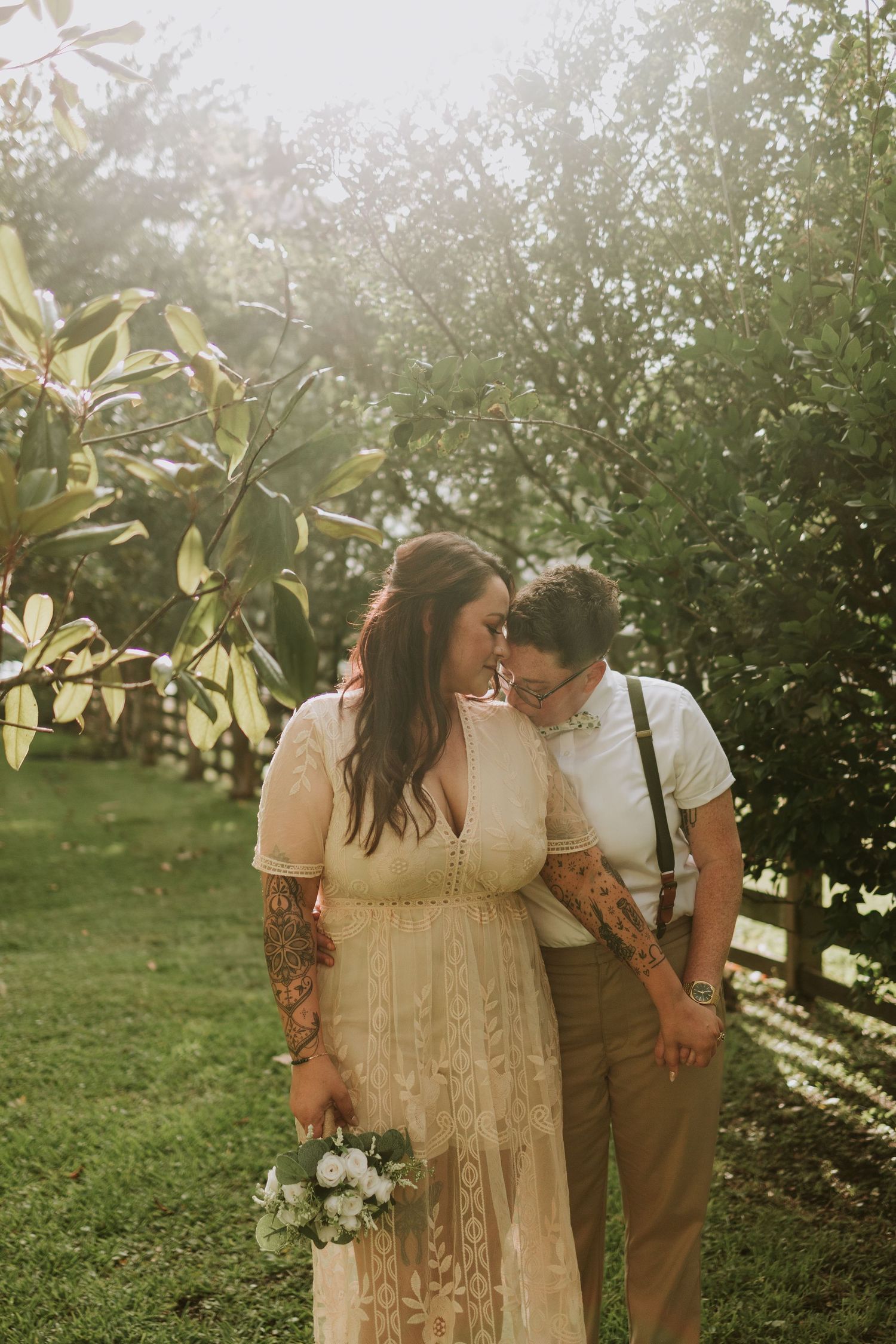 A couple shares a tender forehead touch in a sunlit garden, bride in lace dress holding bouquet, groom in suspenders with tattoos.