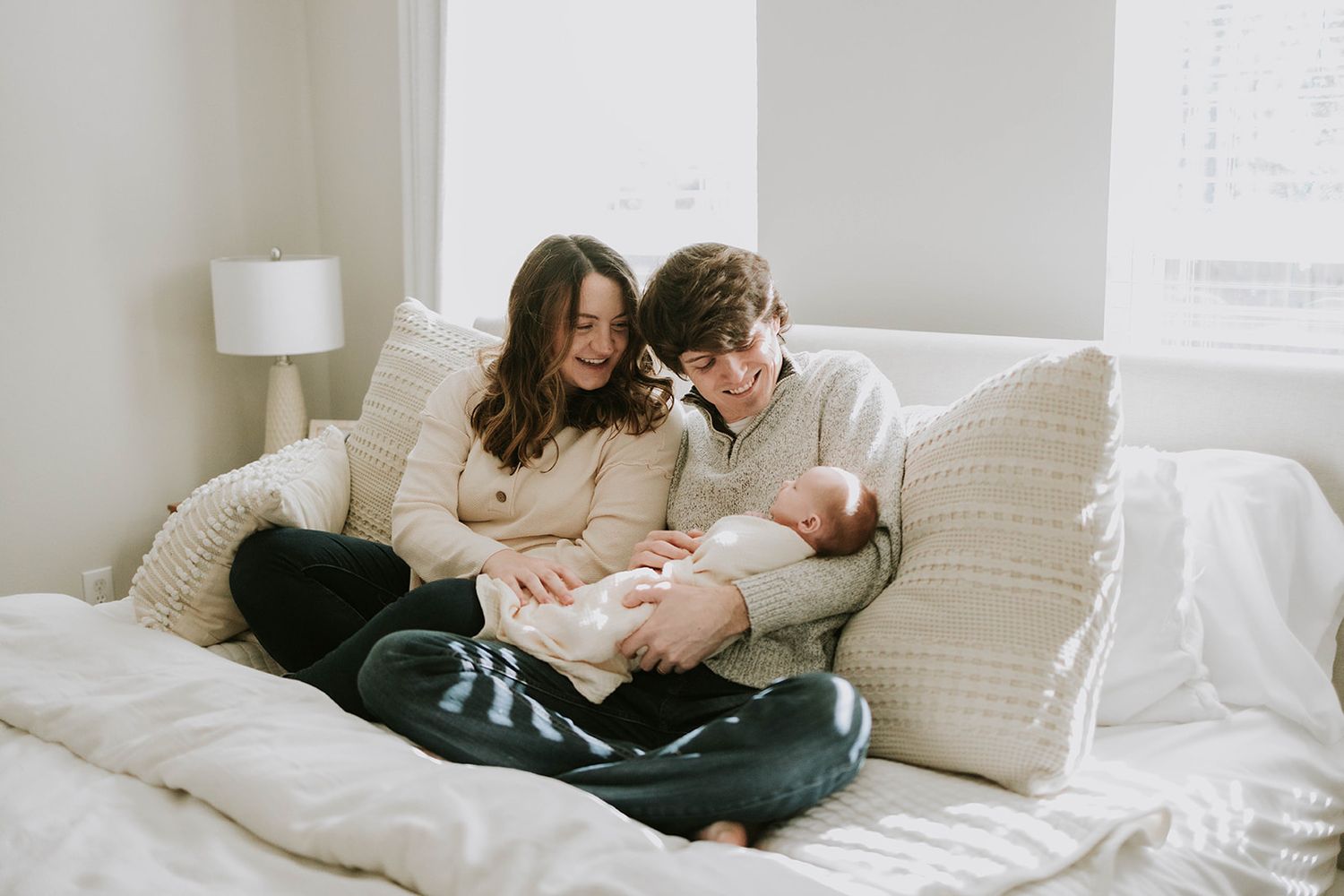 Parents lovingly holding newborn baby while relaxing on white bed.
