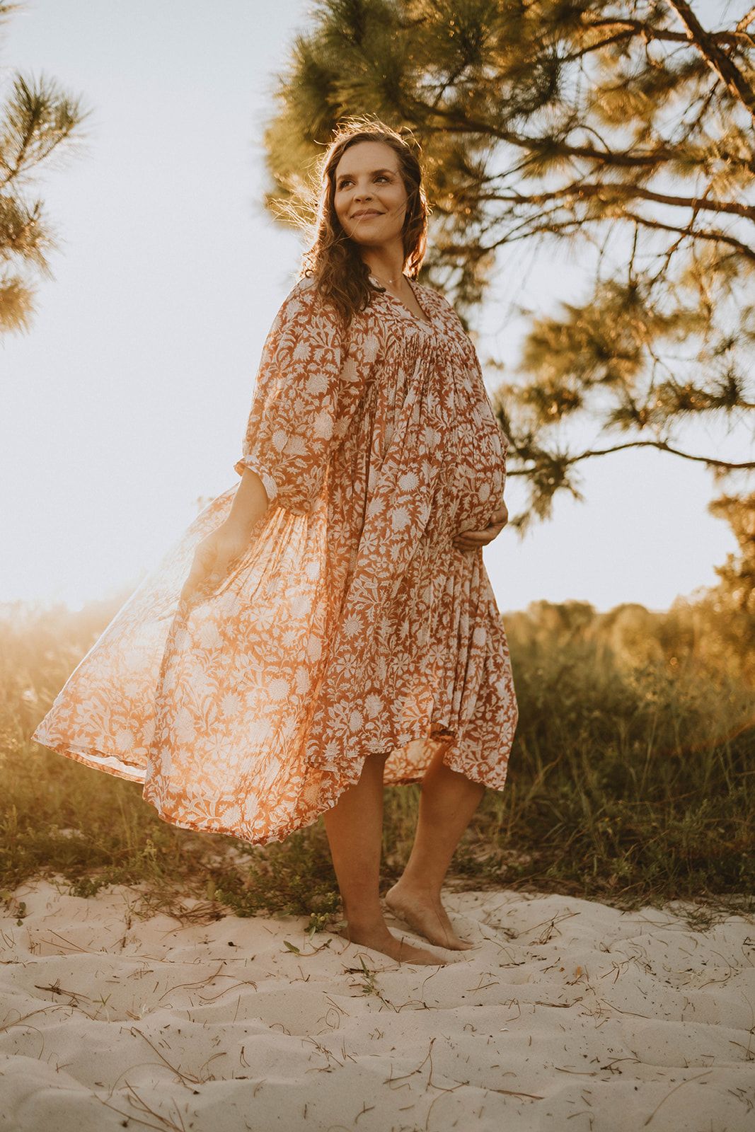 Figure in a floral dress poses barefoot on sandy beach with pine trees at golden hour.
