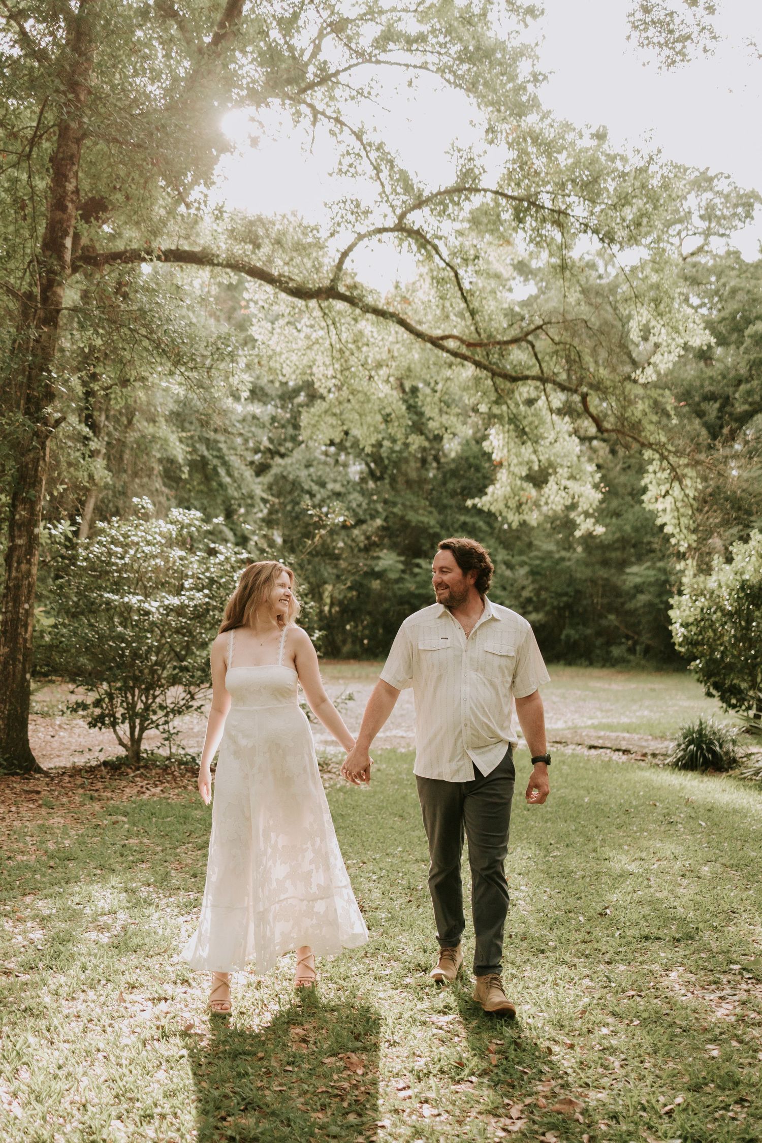 A couple holds hands and walks through a sunlit garden, the woman in a white dress and the man in a light shirt and dark pants.