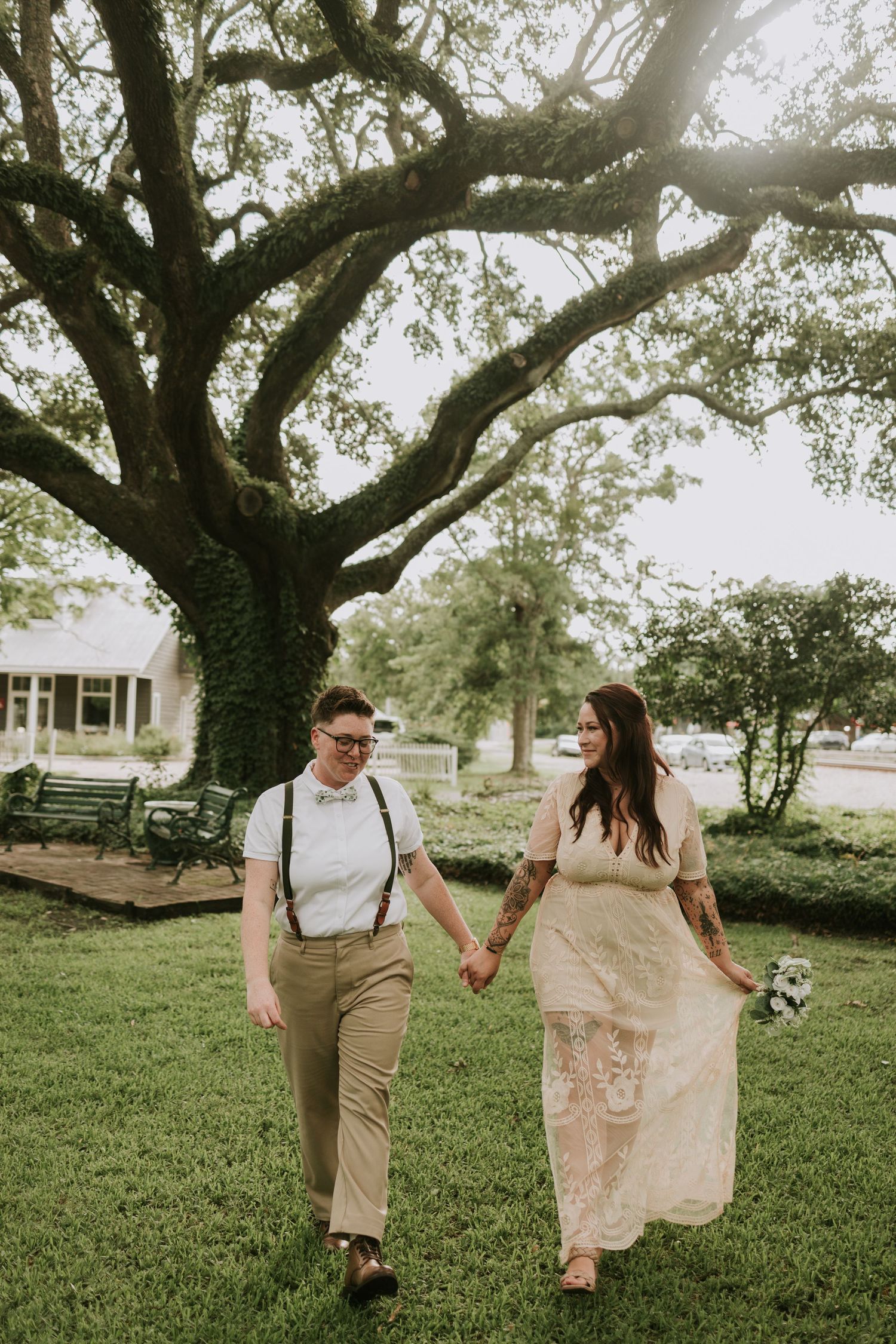 Two brides hold hands walking on green grass beneath a large oak tree on their wedding day.