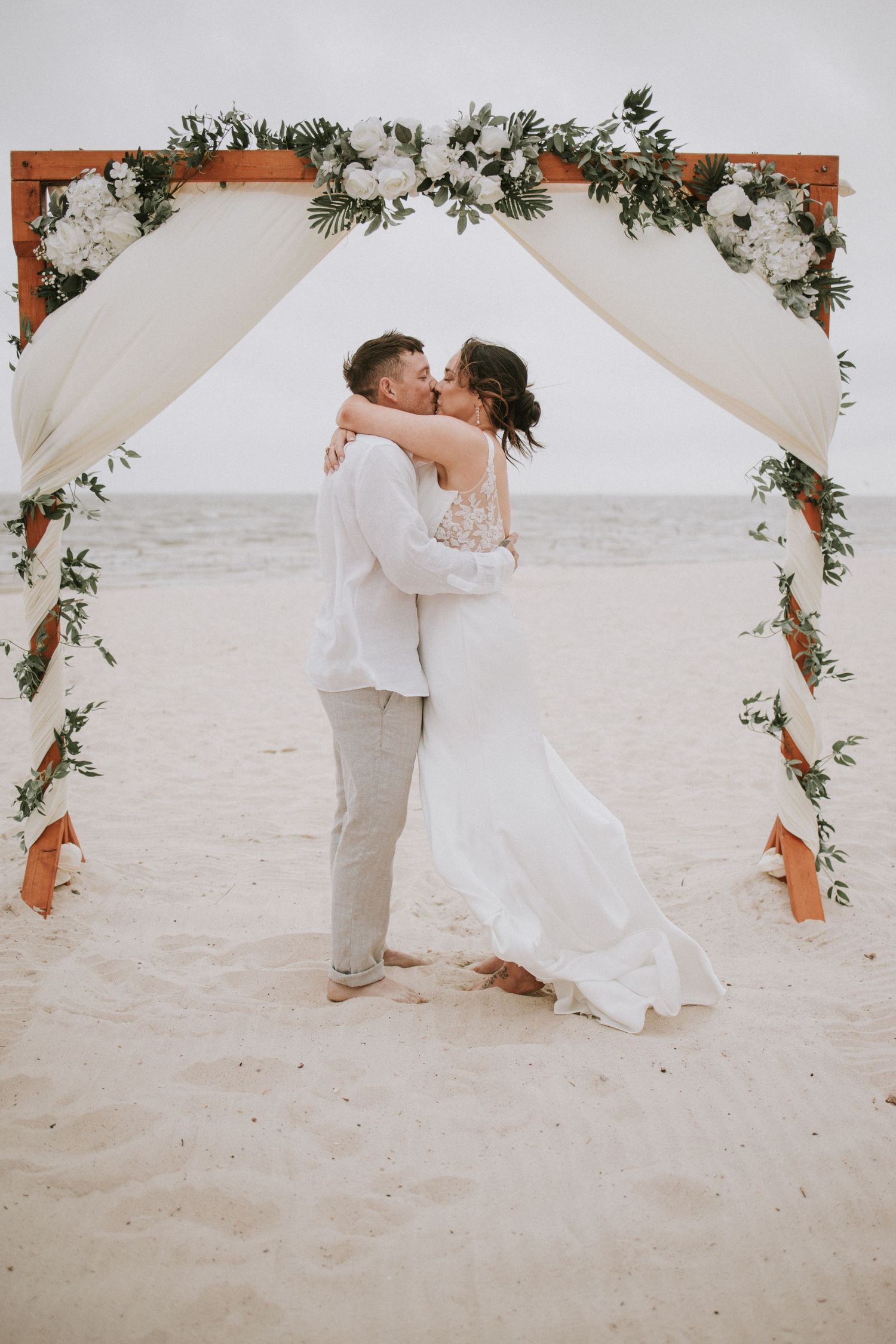 Couple sharing first kiss under floral arch on beach wedding ceremony with white draping and roses.