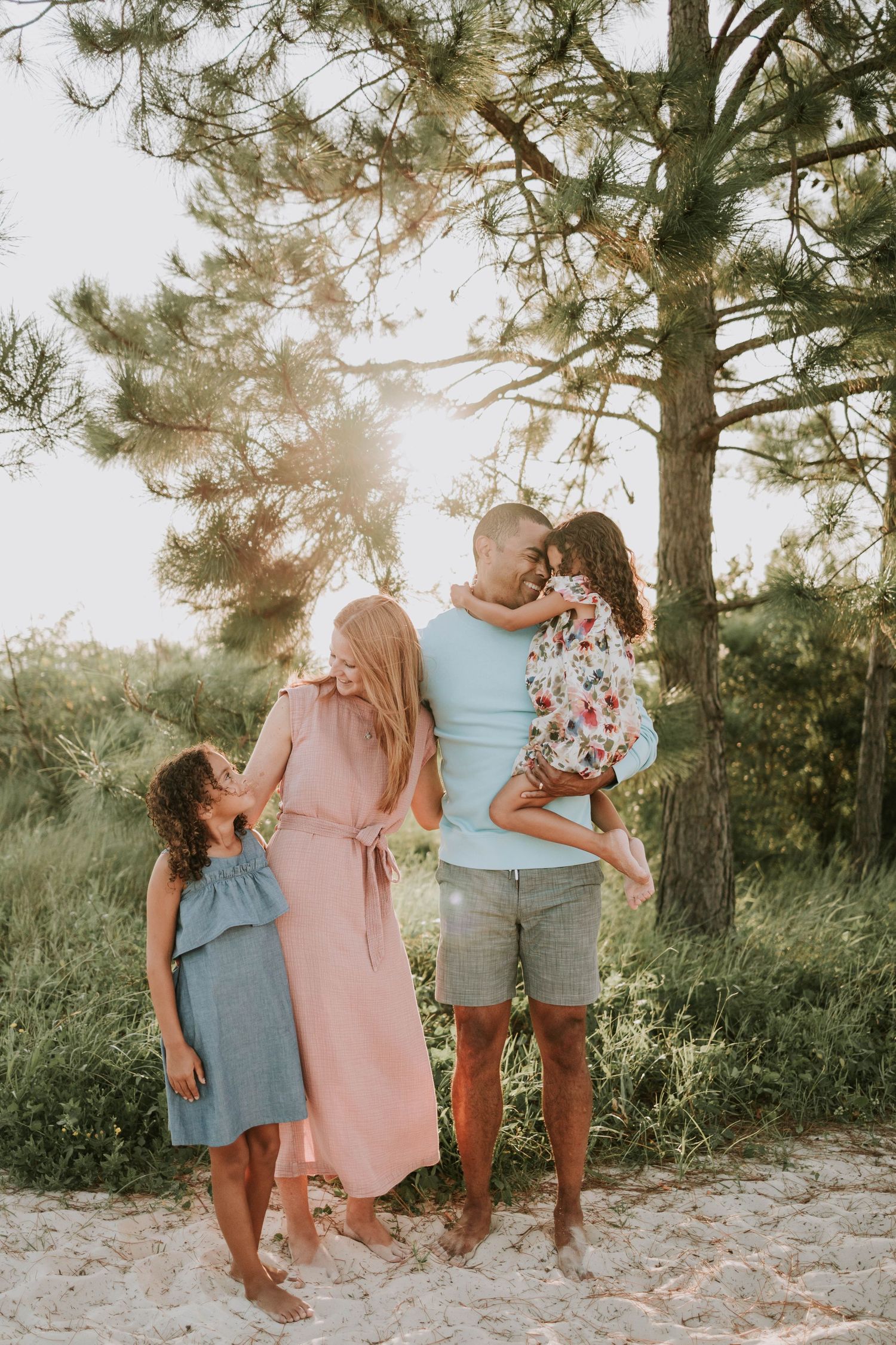 Family of four enjoying a sunny outdoor photo session near pine trees, with parents and two young daughters in pastel outfits.