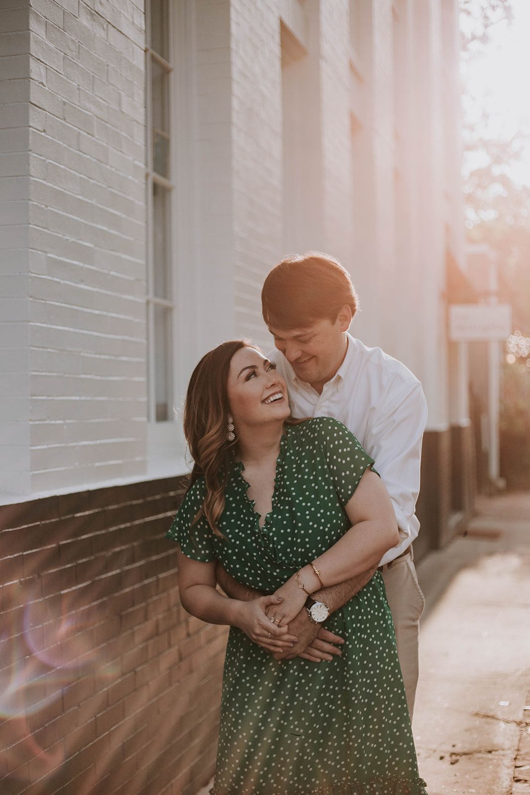 A romantic couple embraces against a white brick wall at sunset, with the woman wearing a green polka dot dress.