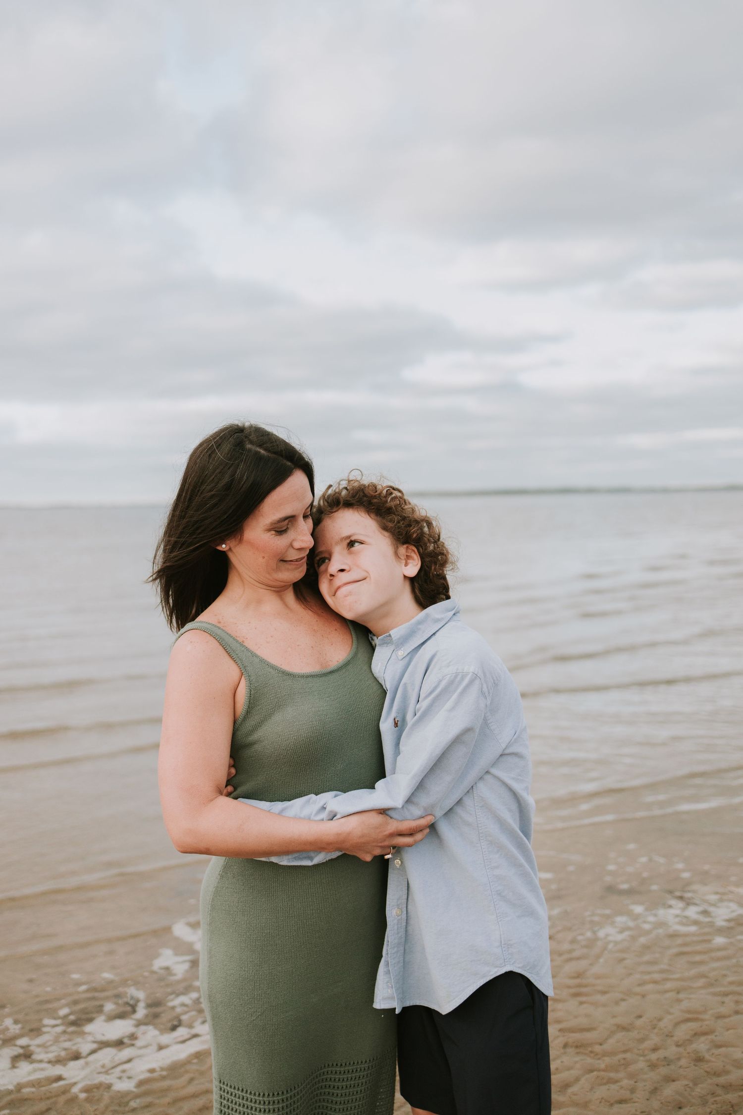 A mother and son share a tender embrace on a sandy beach, with calm ocean waves and cloudy skies behind them.