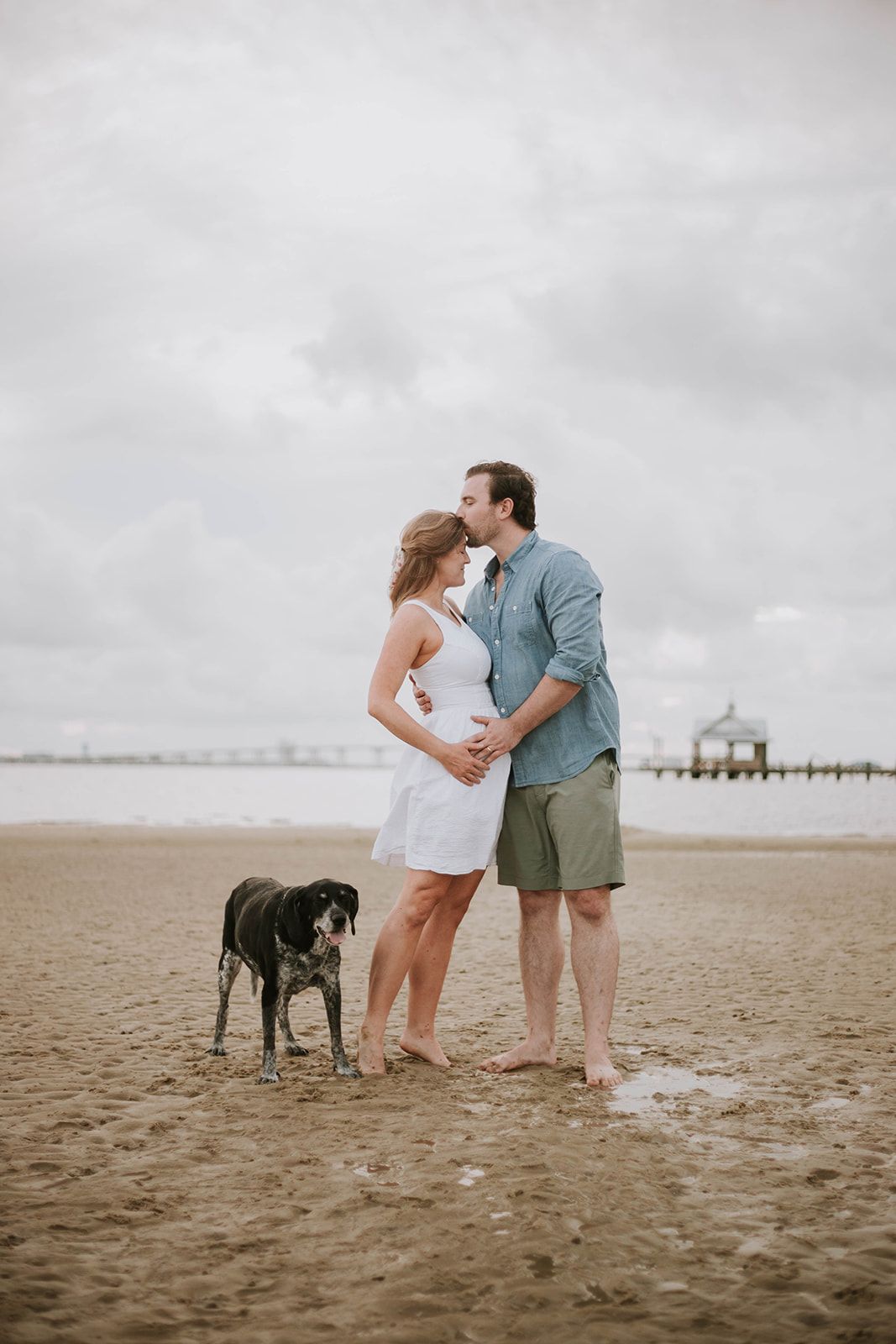 A couple embraces on a beach with their black dog while a lighthouse stands in the distance.