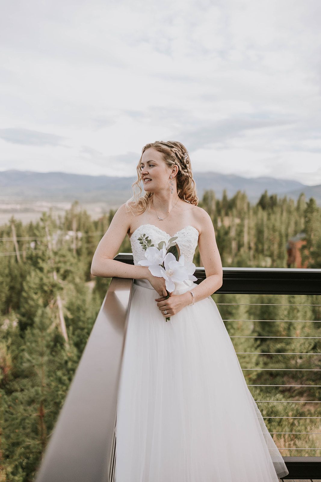 A bride in a flowing white wedding dress laughs joyfully while holding a bouquet against a mountain landscape backdrop.
