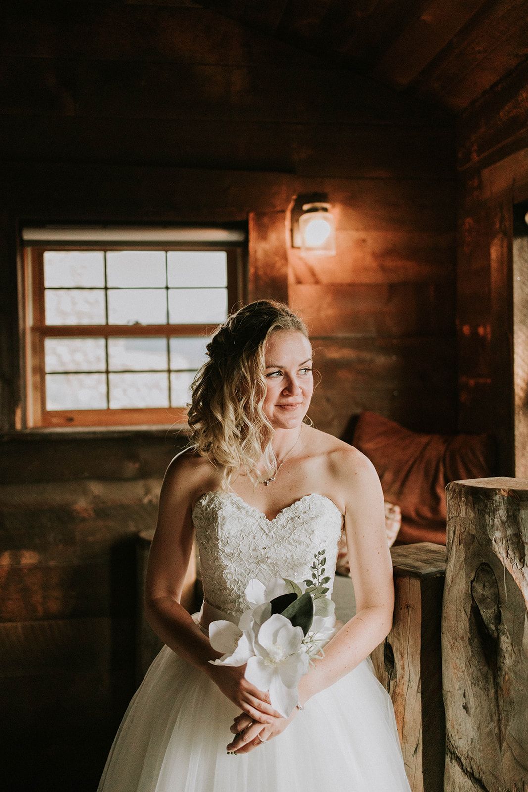 A bride in a white strapless lace wedding gown smiles while standing in a rustic cabin with warm lighting.
