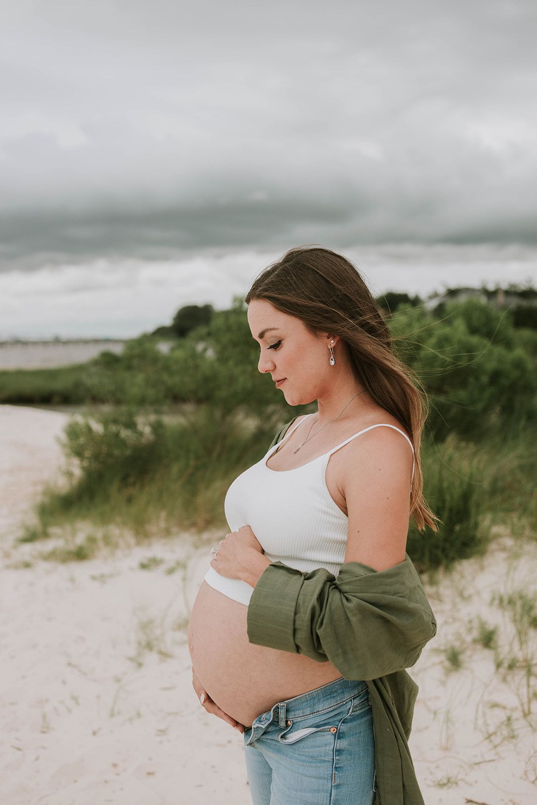 Side view of expectant mother in white tank top and green jacket on beach.