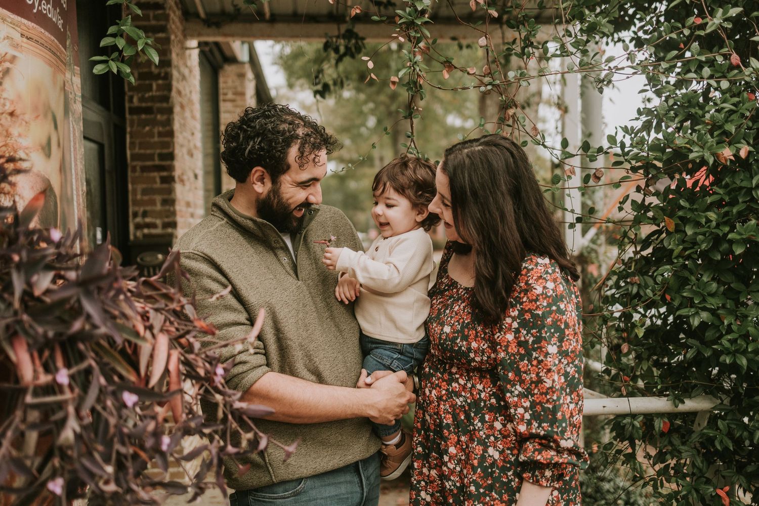 A young family with a bearded father and brunette mother smiling at their toddler outdoors near lush greenery.