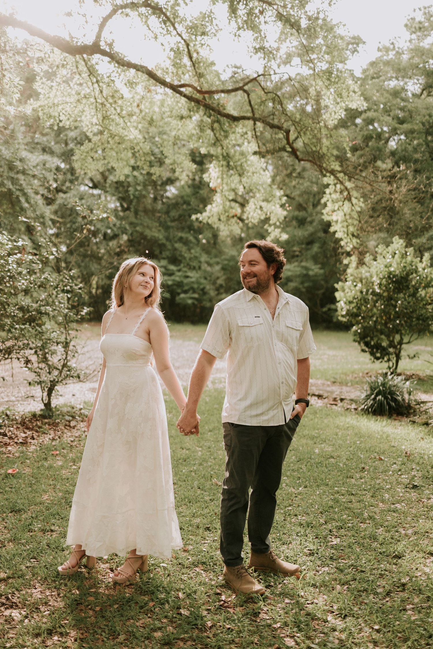 A couple holds hands and walks through a lush green park, the woman in a white dress and the man in a light shirt.