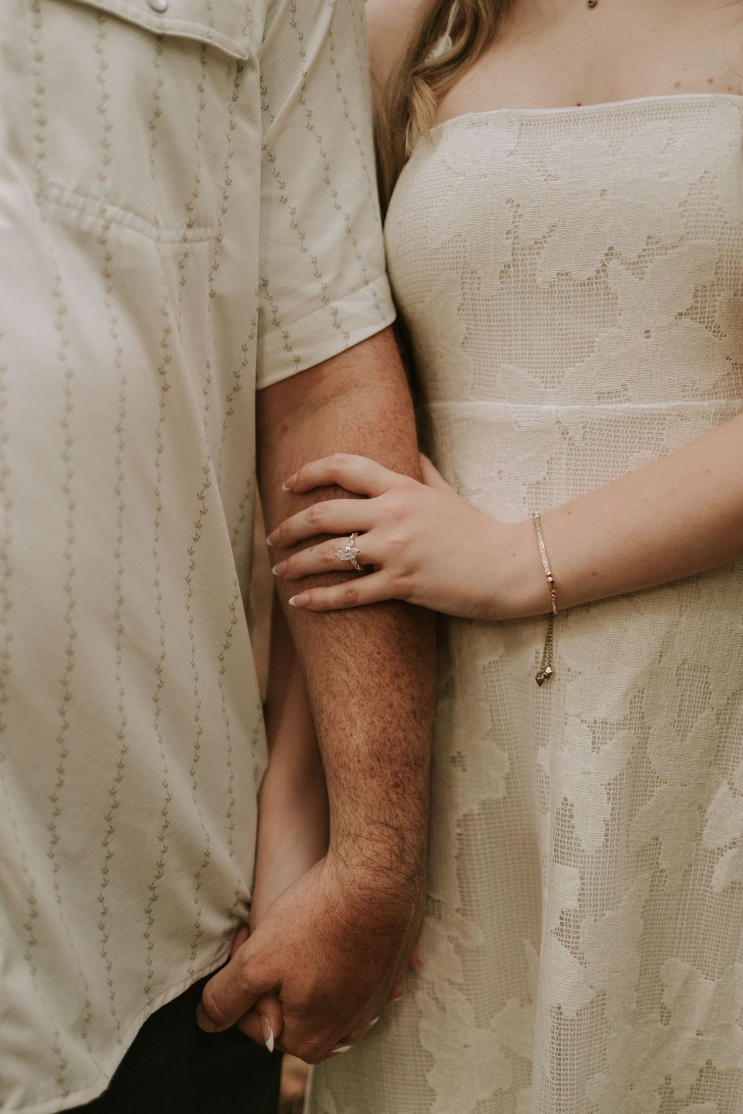 Close-up of a couple holding hands, bride in lace dress with ring and bracelet, groom in cream shirt.