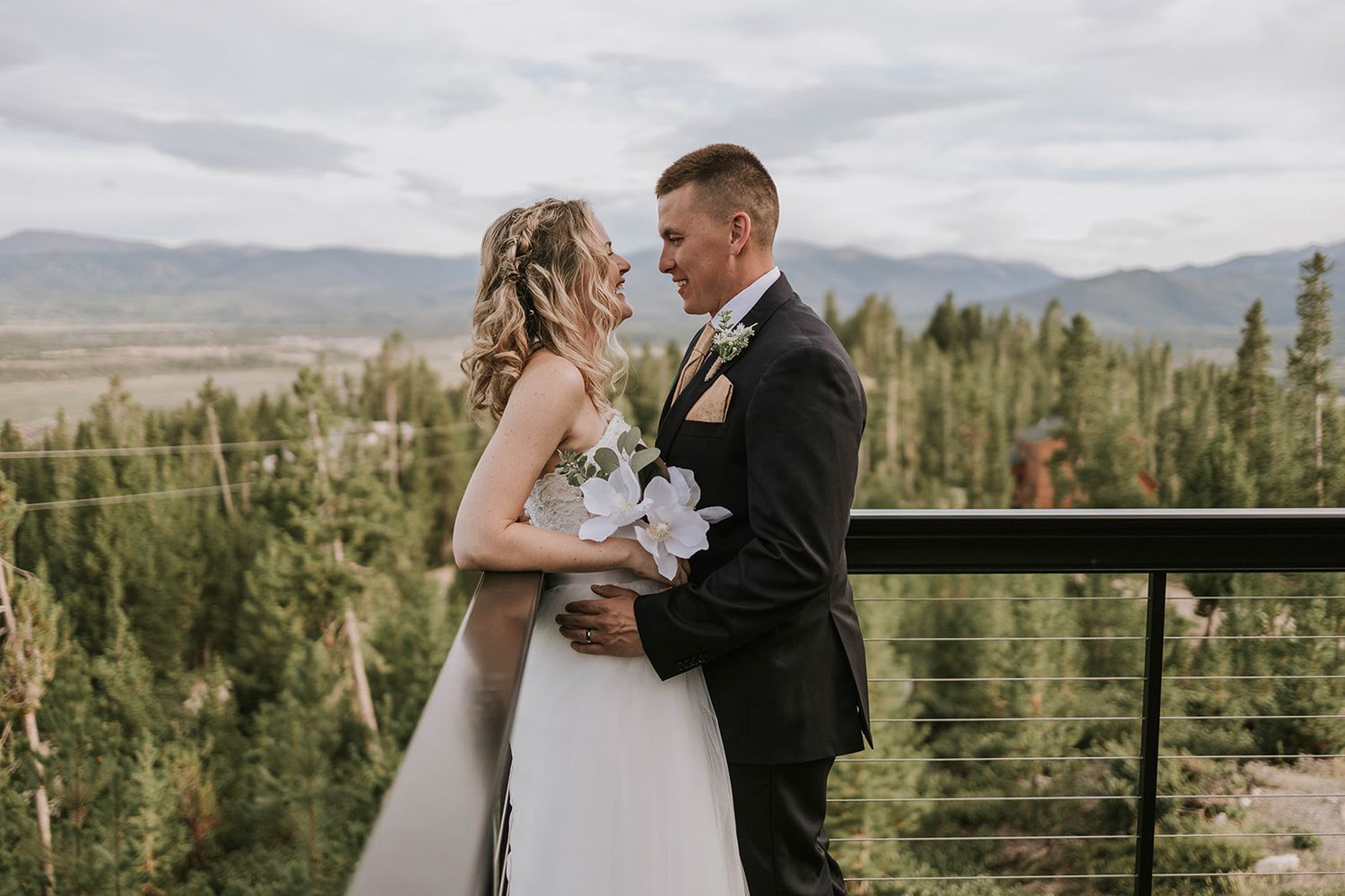 Couple shares romantic moment at mountain overlook with evergreen forest and dramatic cloudy sky in background.