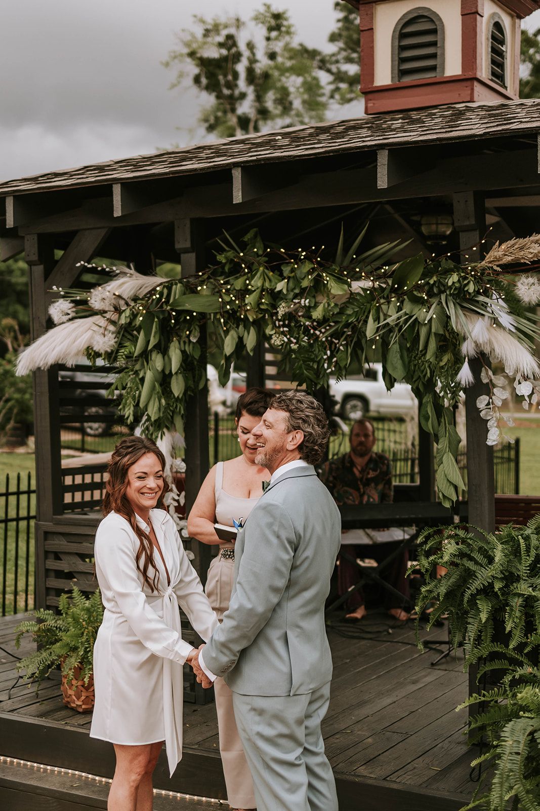 Couple stands together under a rustic gazebo decorated with greenery and white florals during an intimate wedding ceremony.